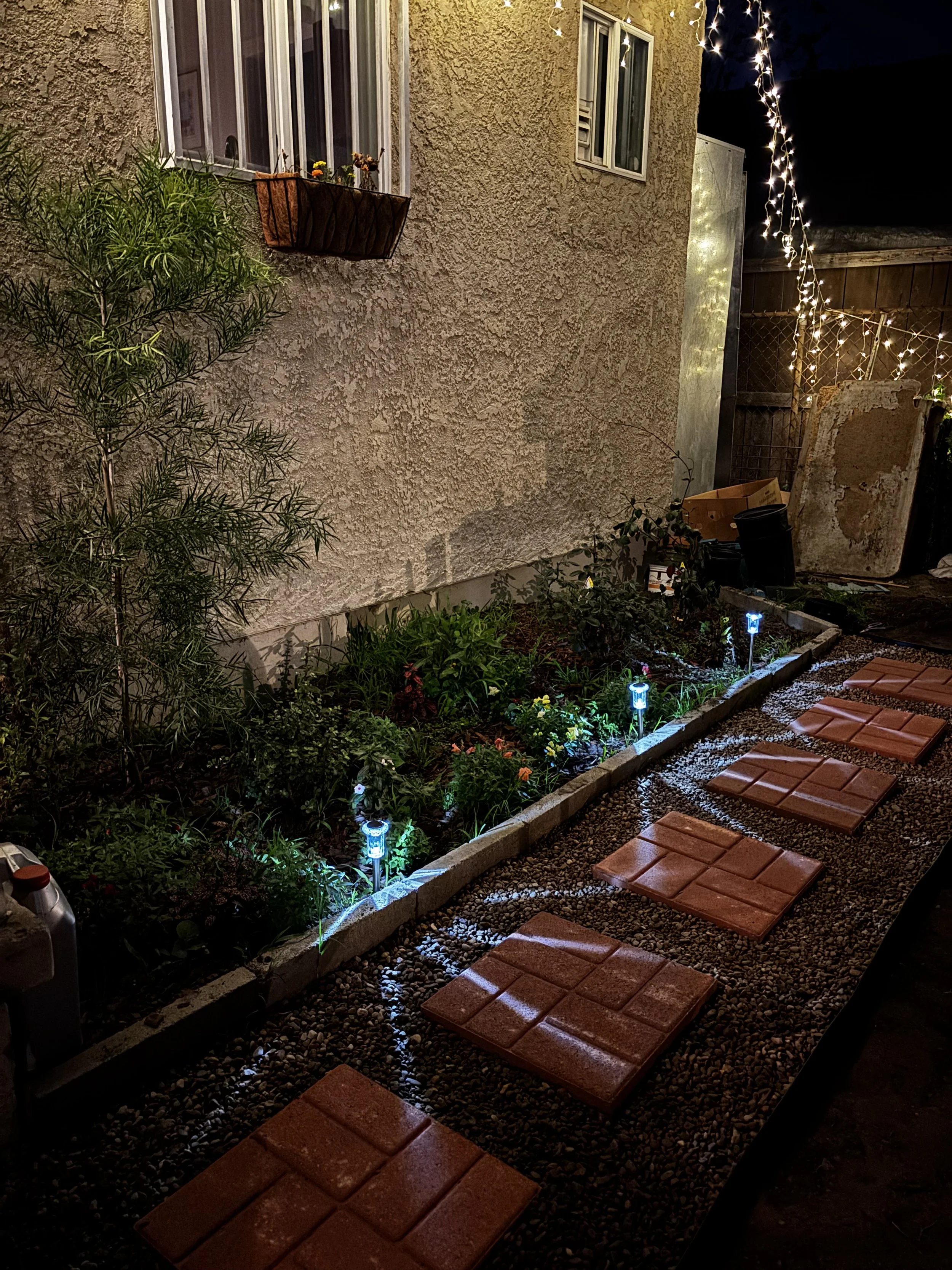 Nighttime view of a small garden bed with illuminated pathway markers, decorative string lights on the wall, a window with flower box plants, and a wooden fence in the background.