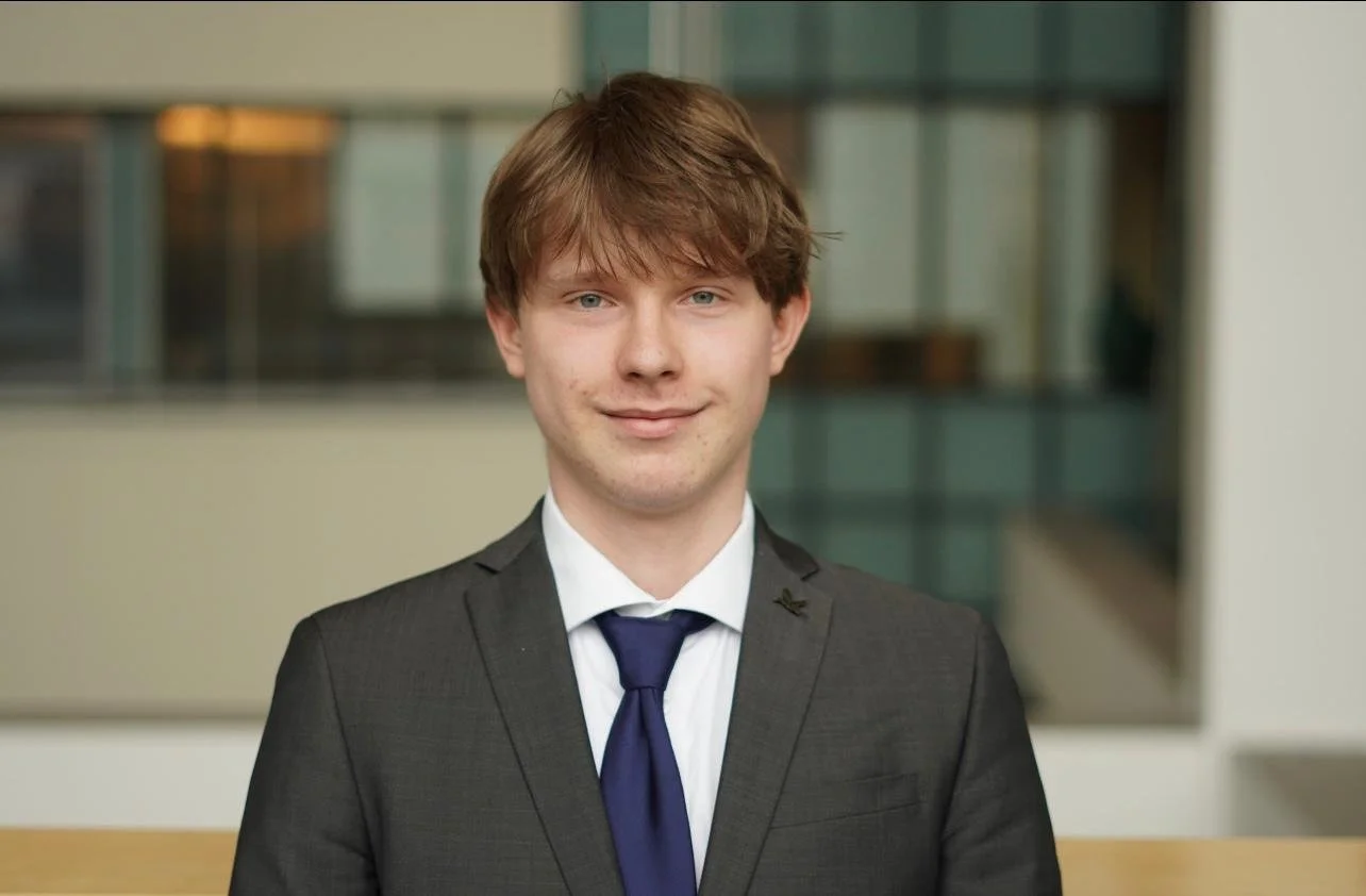A young man with light brown hair wearing a dark gray suit, a light blue shirt, and a plaid tie, standing outdoors in front of a glass building.