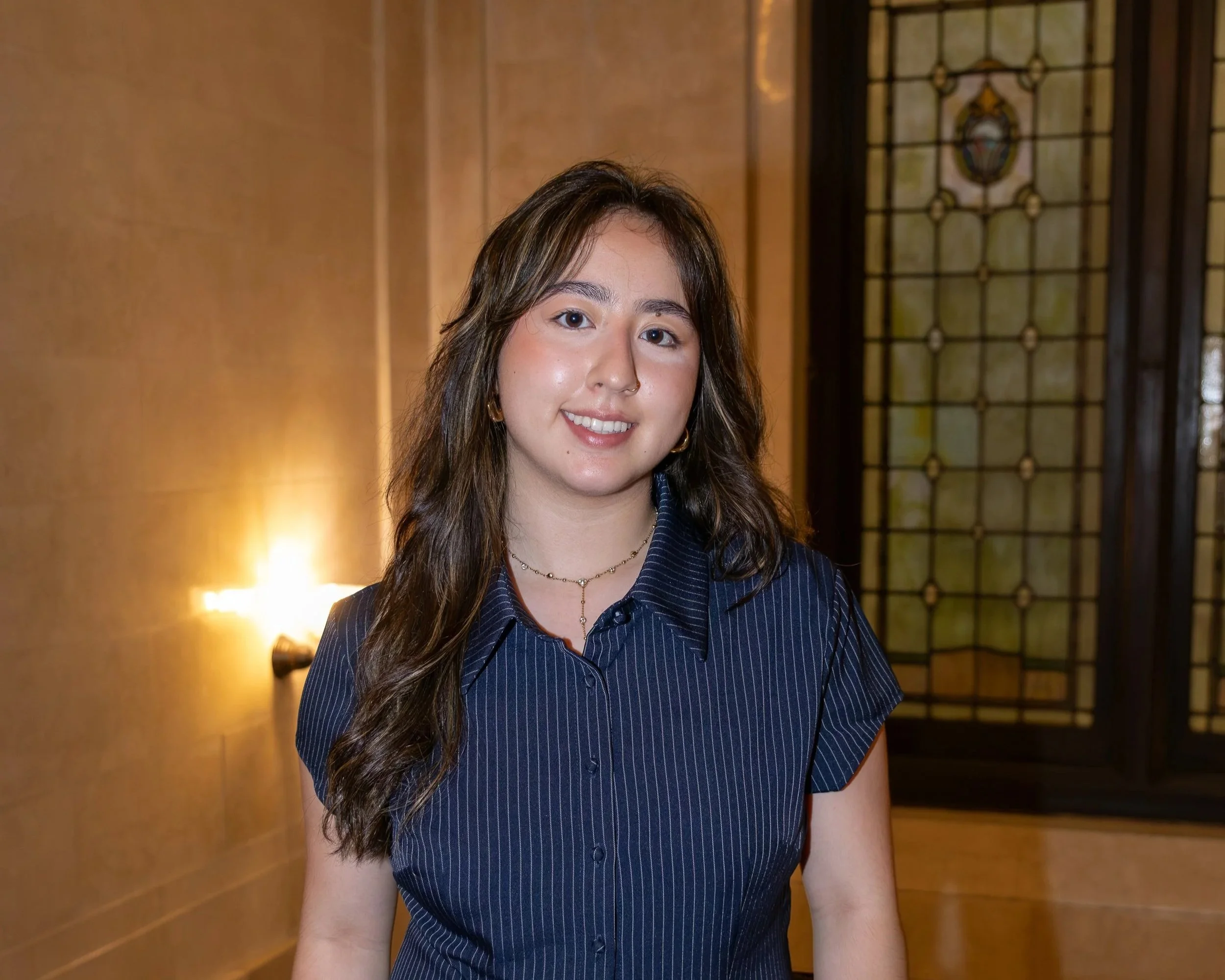 A young woman with long dark hair, wearing a black blazer, white shirt, and black tie, smiling in an indoor setting with background blurred out.