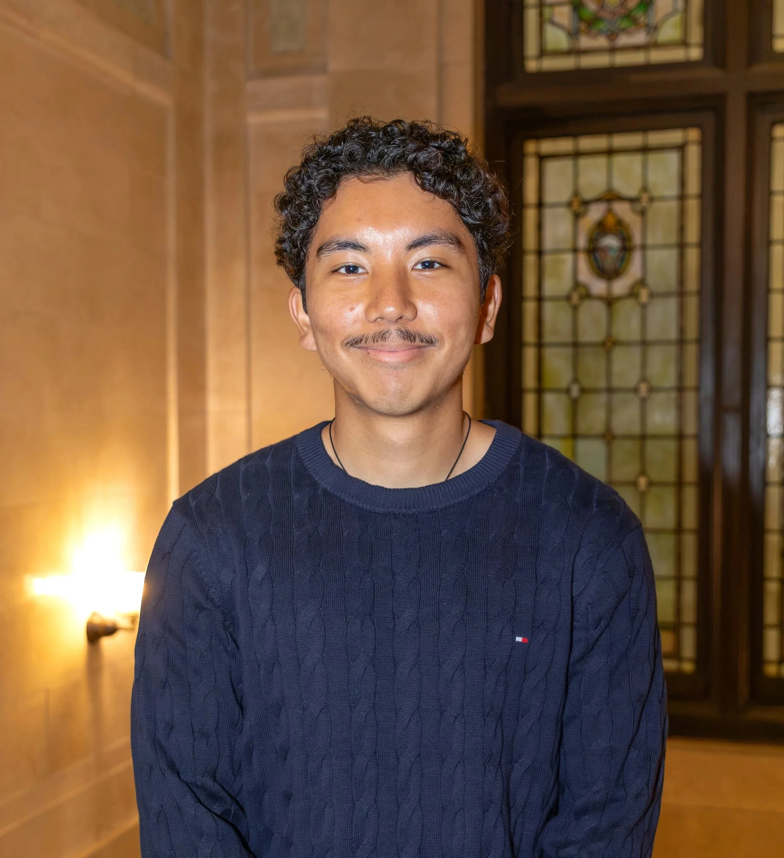 A young man in a black suit and white shirt smiling in an indoor setting with blurred background.