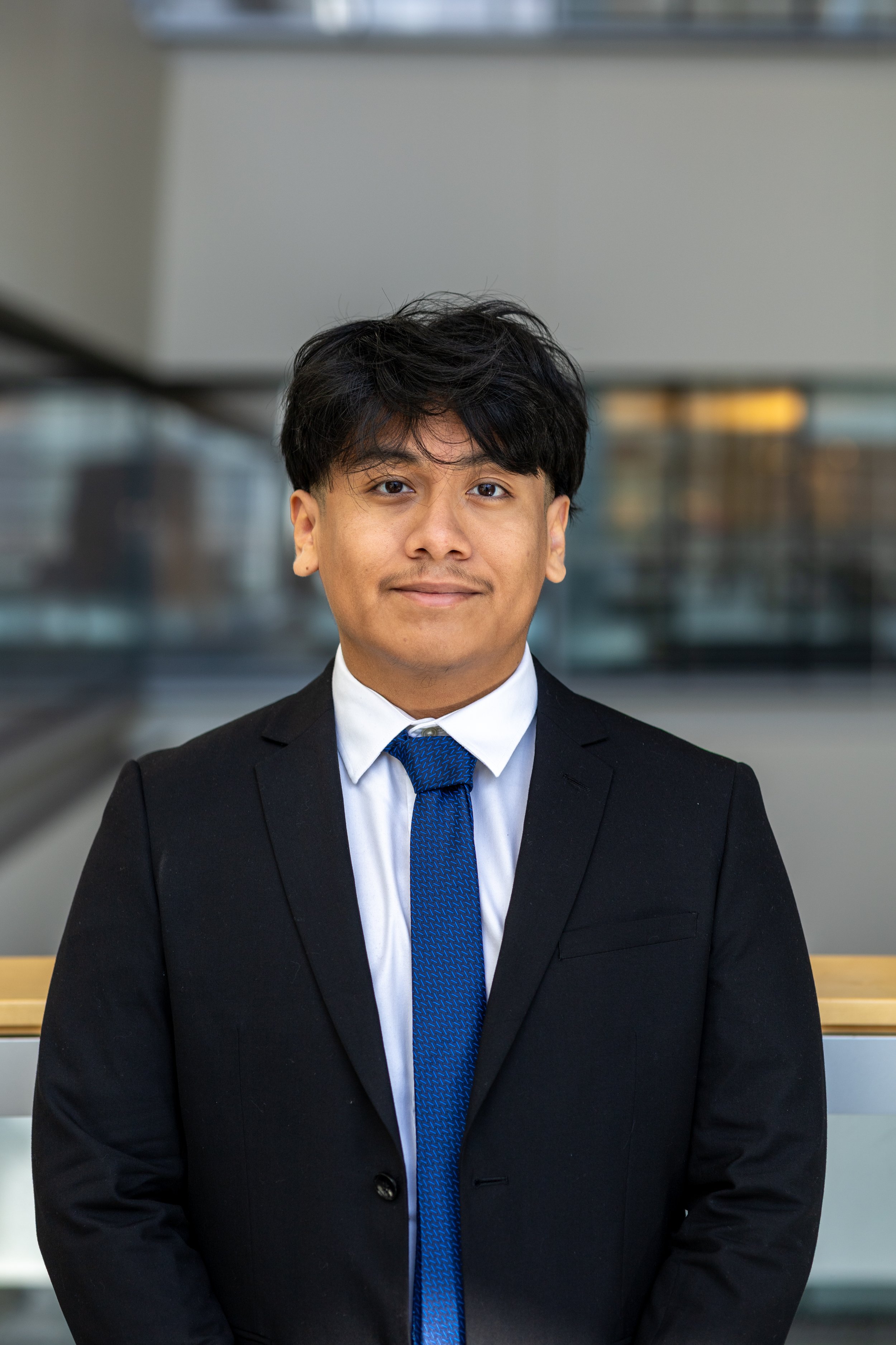 Portrait of a young woman with long brown hair, wearing a black blazer and white shirt, standing indoors with a glass wall in the background.