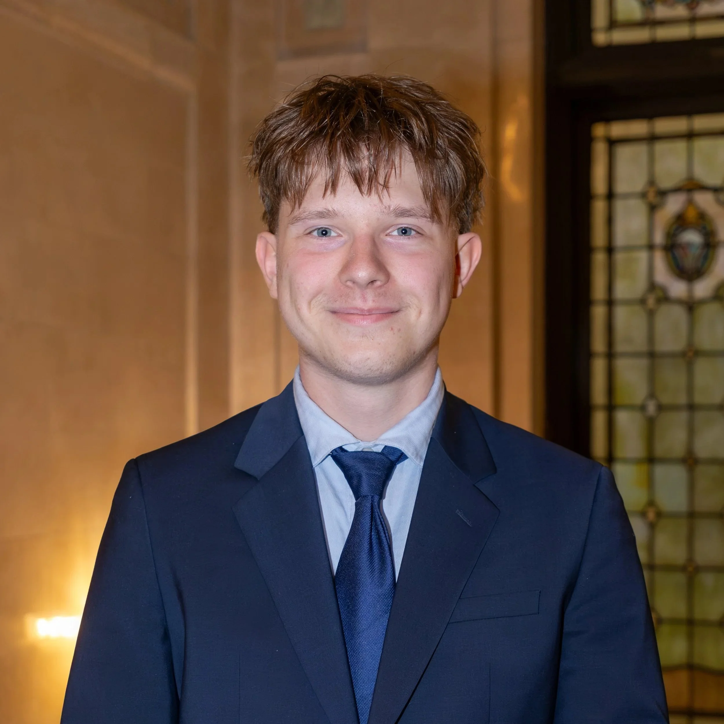 A young man with light brown hair wearing a dark gray suit, a light blue shirt, and a plaid tie, standing outdoors in front of a glass building.