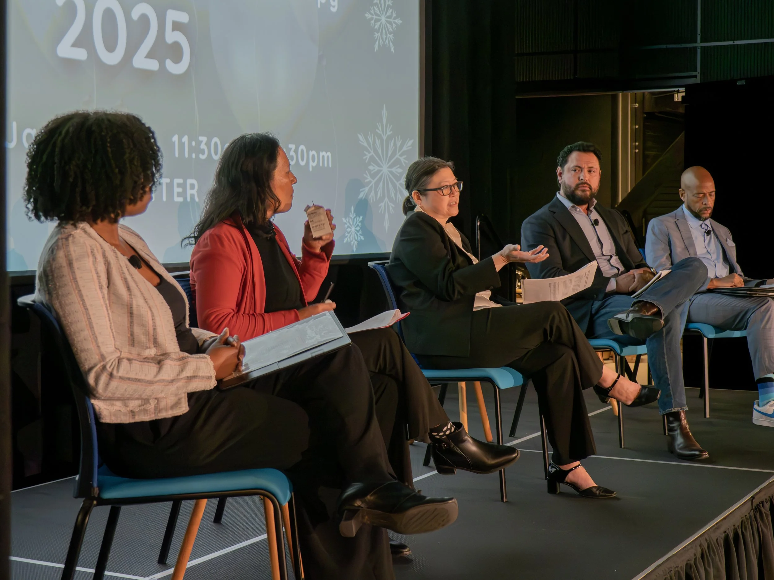 A panel of five diverse individuals seated on stage during a conference or discussion, with a projected screen behind them displaying event details and holiday-themed graphics.