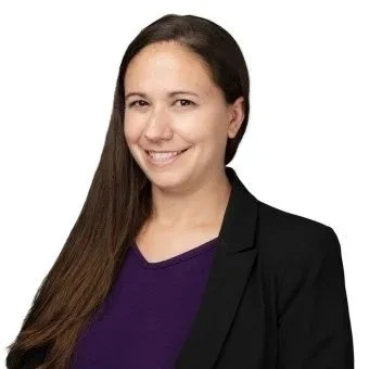 A woman with long brown hair smiling, wearing a purple top and black blazer against a white background.