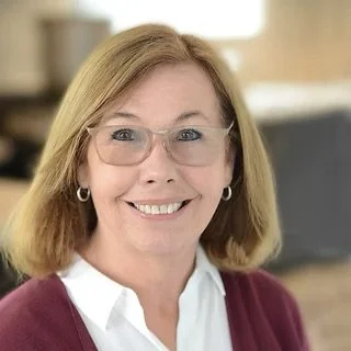 Portrait of a smiling woman with shoulder-length blonde hair wearing glasses, a white shirt, and a maroon sweater, indoors.