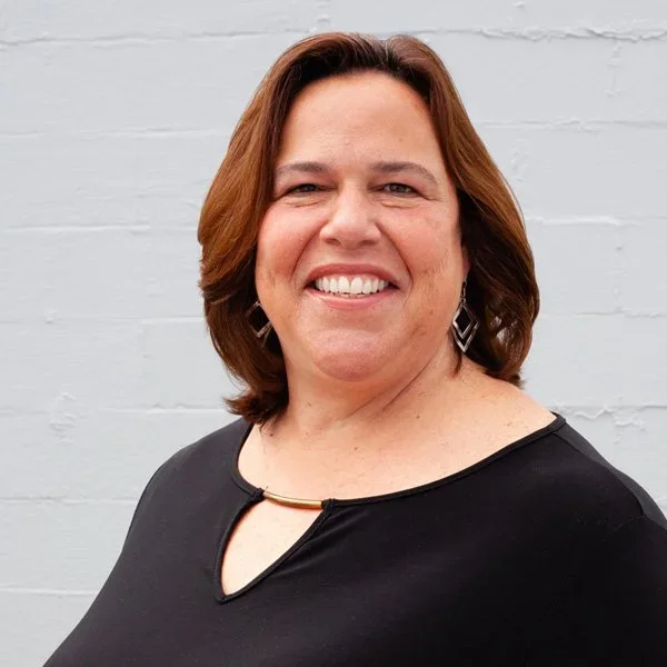 A woman with shoulder-length brown hair smiling, wearing a black top with a small cutout and gold jewelry, standing against a plain white brick wall.