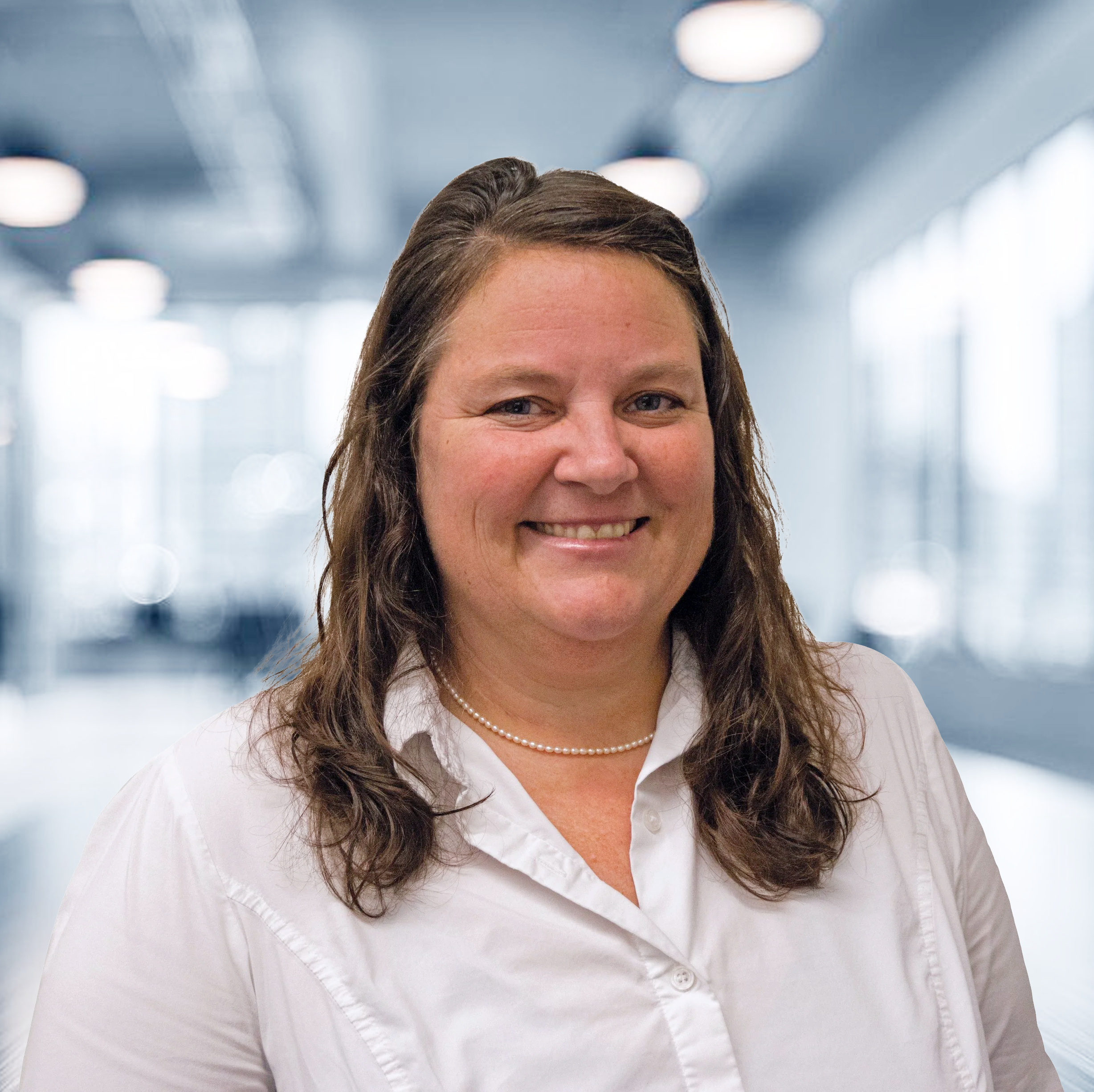 A woman with shoulder-length brown hair, wearing a white blouse and a pearl necklace, smiling at the camera in a brightly lit modern indoor setting.