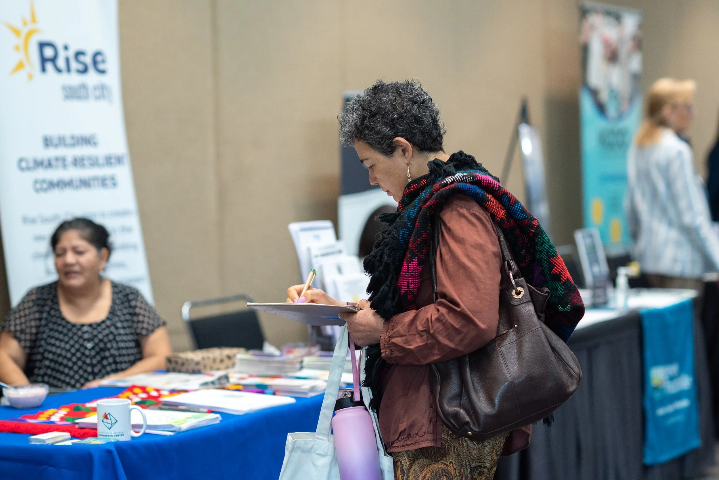 A woman with short curly hair, wearing a brown jacket and colorful scarf, is signing a book or document at a booth during an event. The booth has a blue tablecloth with various items, including a white mug with a logo, displayed on it. Background features a woman sitting at the booth and banners with text.