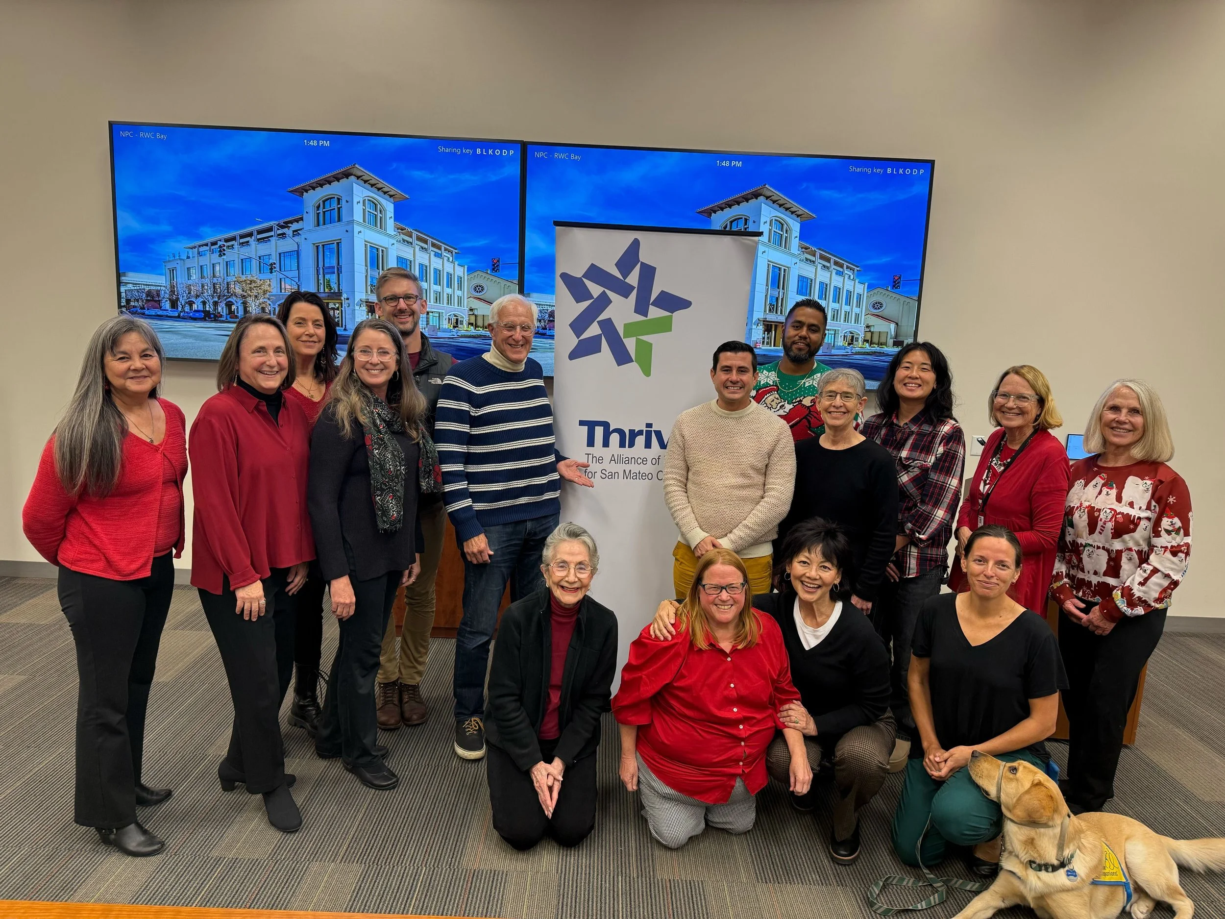 A group of 16 people, including a guide dog, posing in a conference room with a banner that says 'Thrive, The Alliance for San Mateo C'. Behind them on the wall are two large screens displaying a building with a blue sky. The individuals are dressed in casual and holiday-themed attire, smiling at the camera.
