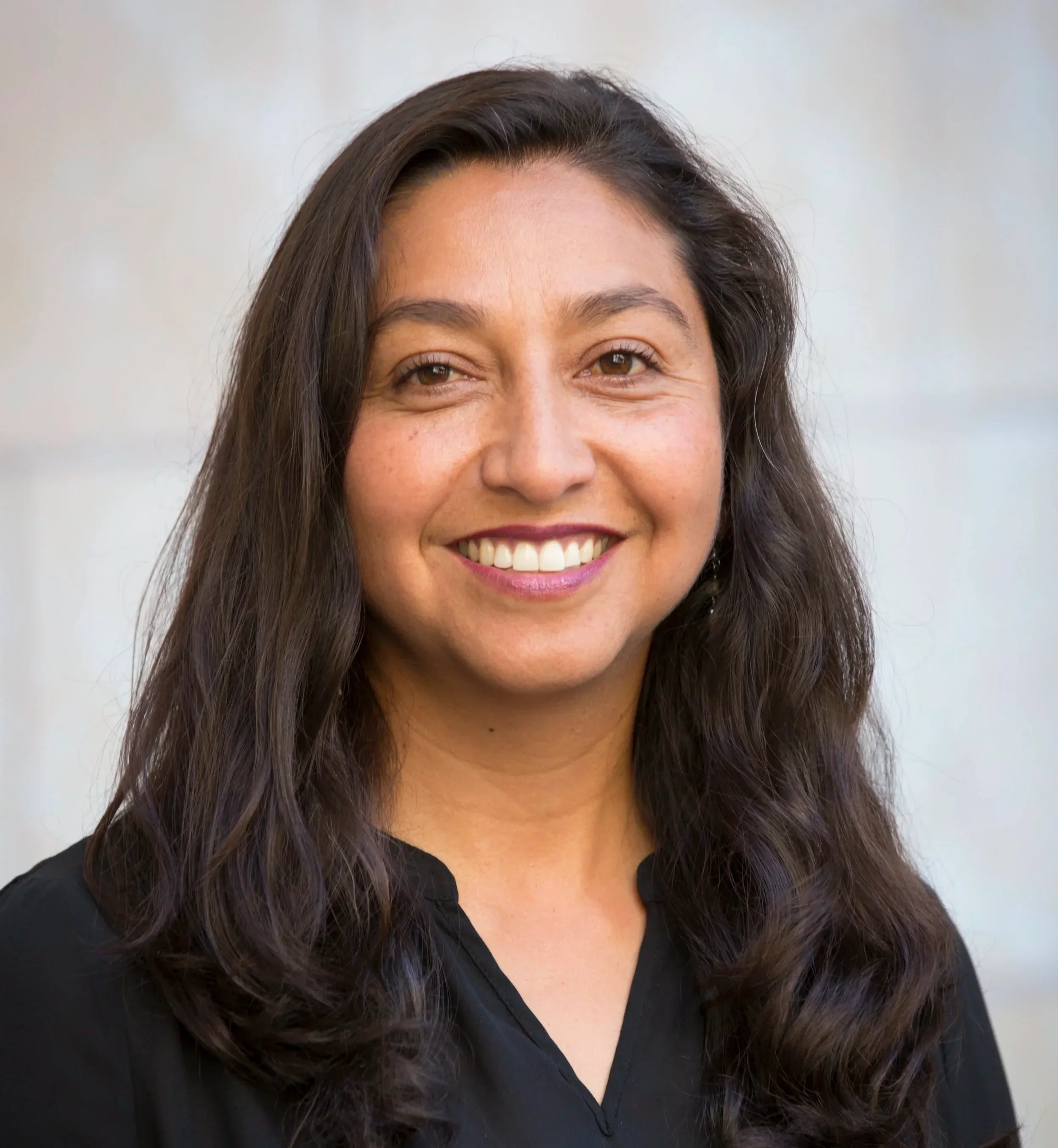 A woman with long dark wavy hair and a big smile, wearing a black top.