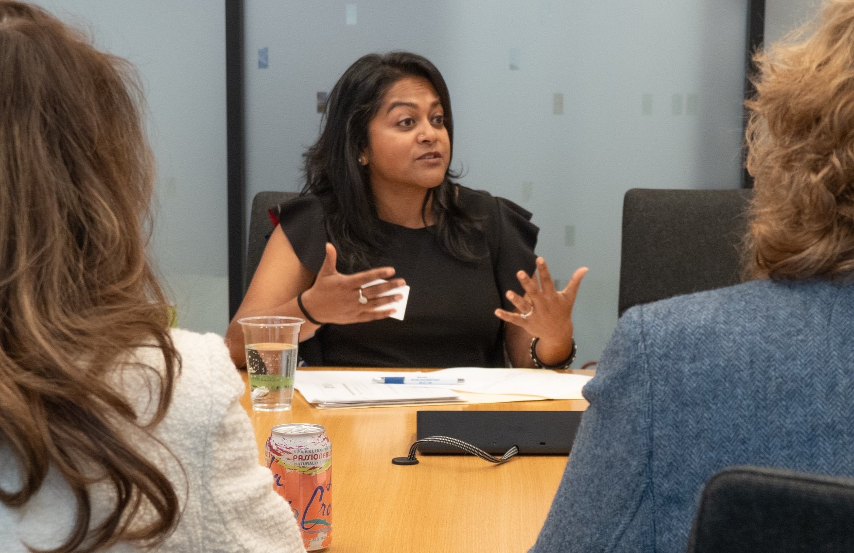 A woman is sitting at a conference table speaking and gesturing with her hands; three people are partially visible around her.