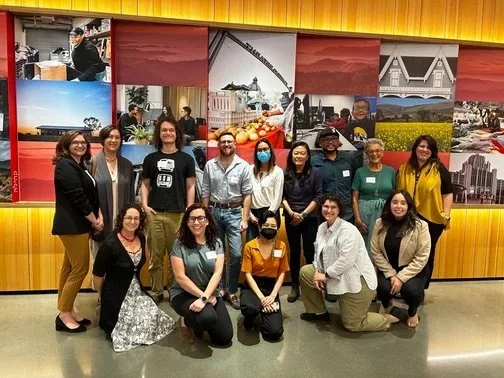 Group of twelve diverse people posing indoors in front of a colorful mural featuring cityscape and architectural images.