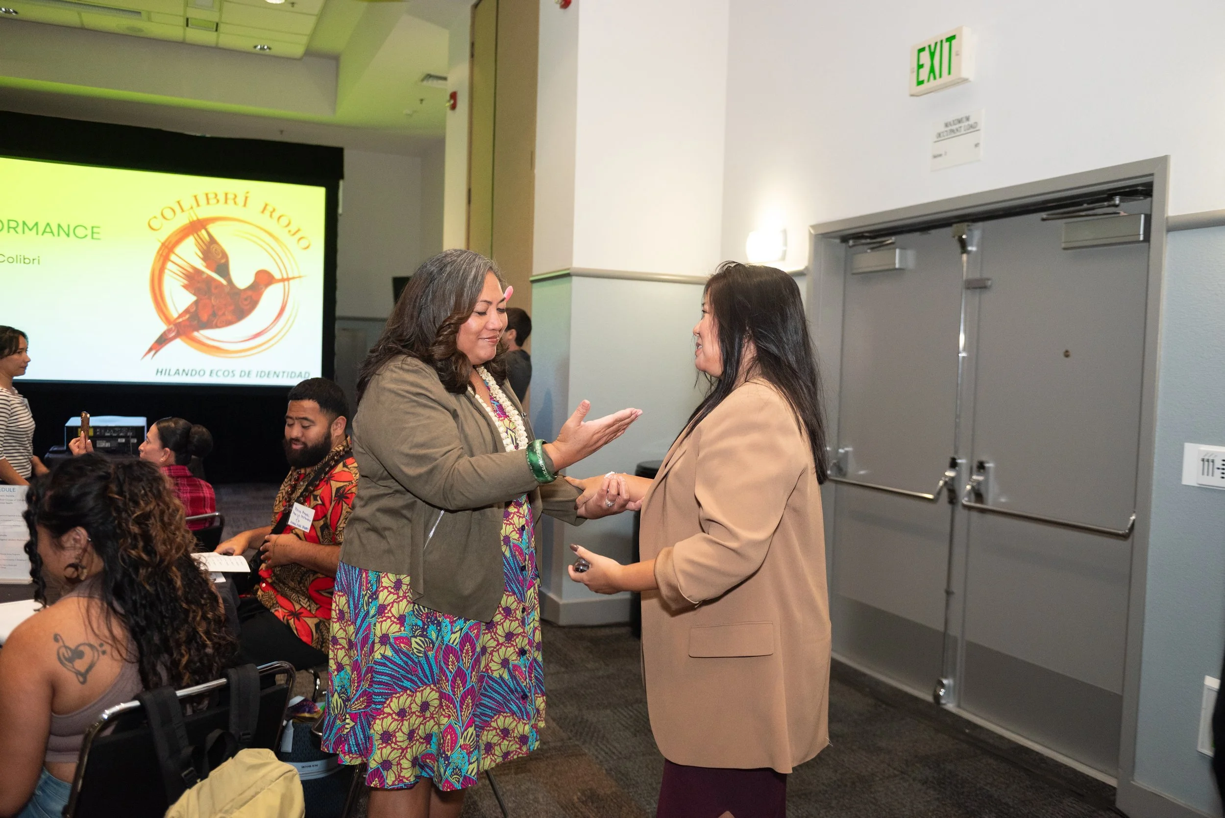 Two women shaking hands and talking in a conference room with seated attendees and a large screen showing a logo with a hummingbird and the text 'Colibrí Rojo' in the background.