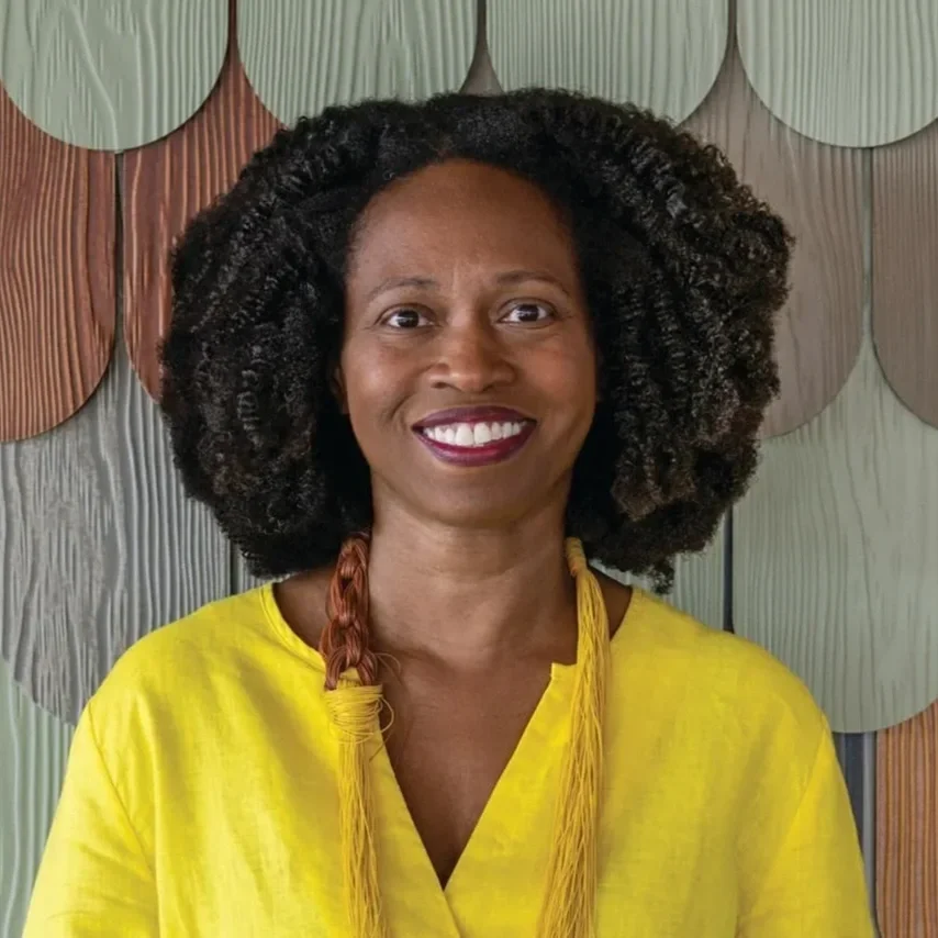 A woman with dark curly hair smiling, wearing a yellow top and standing in front of a colorful wooden wall.