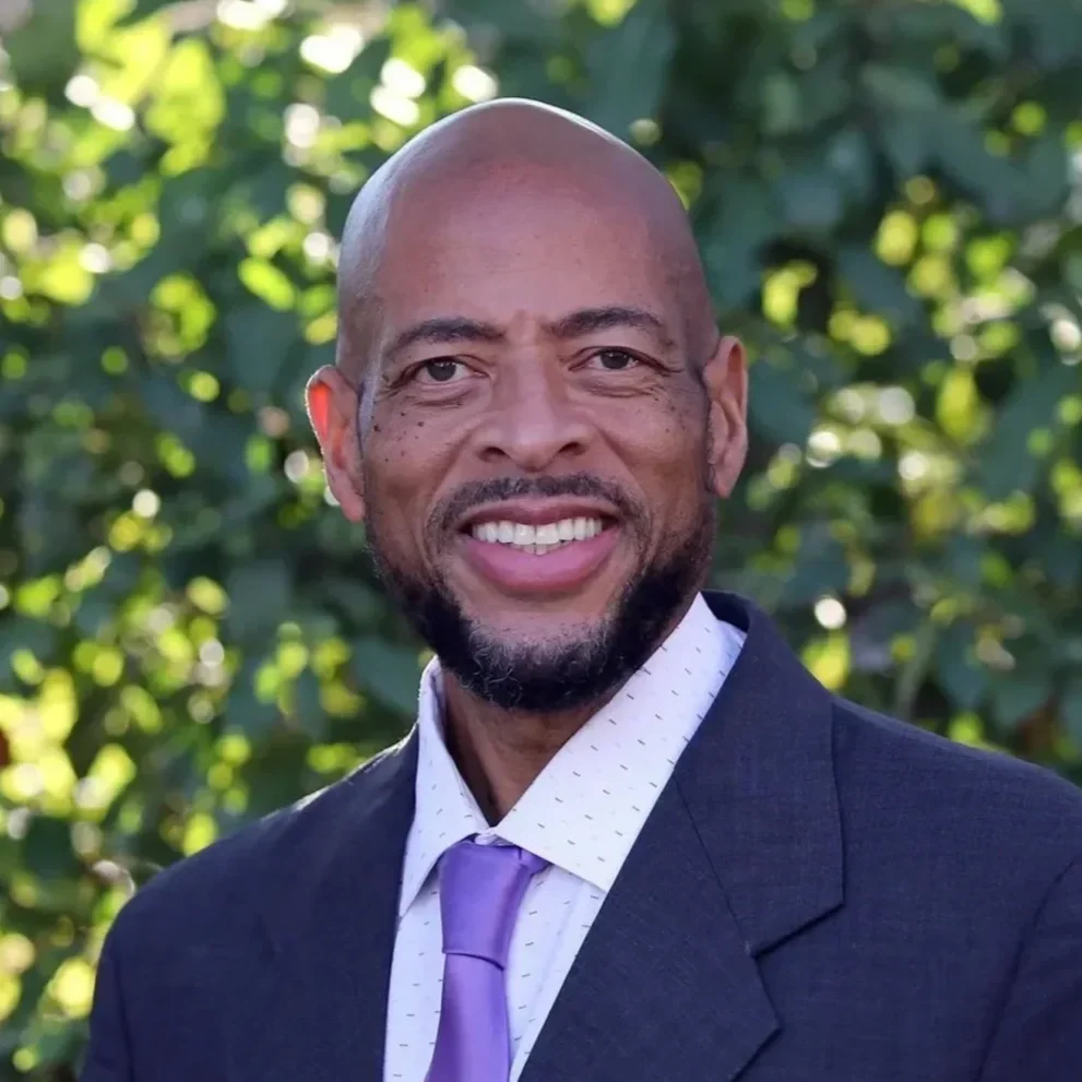 A smiling man wearing a suit jacket, white shirt, and purple tie, standing outdoors with greenery in the background.