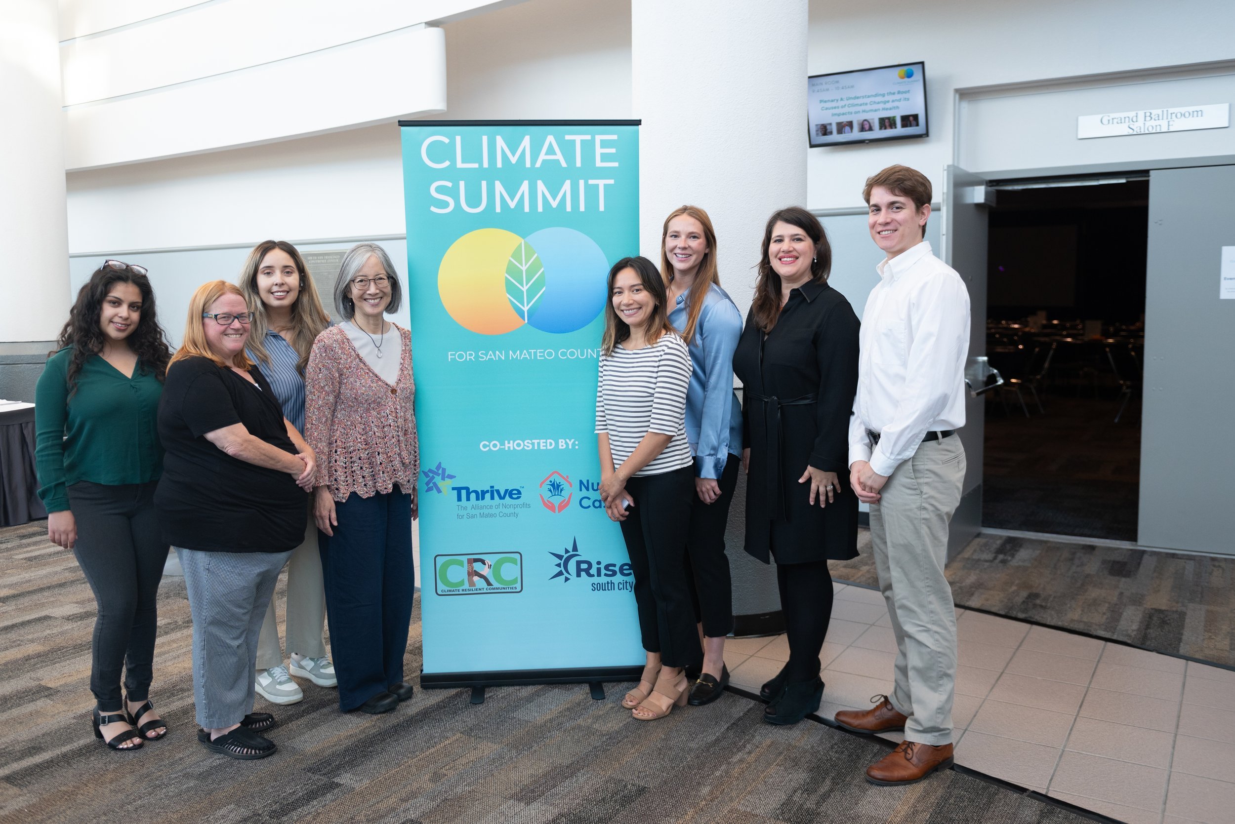 Group of eight people standing next to a 'Climate Summit' banner in an indoor conference area.