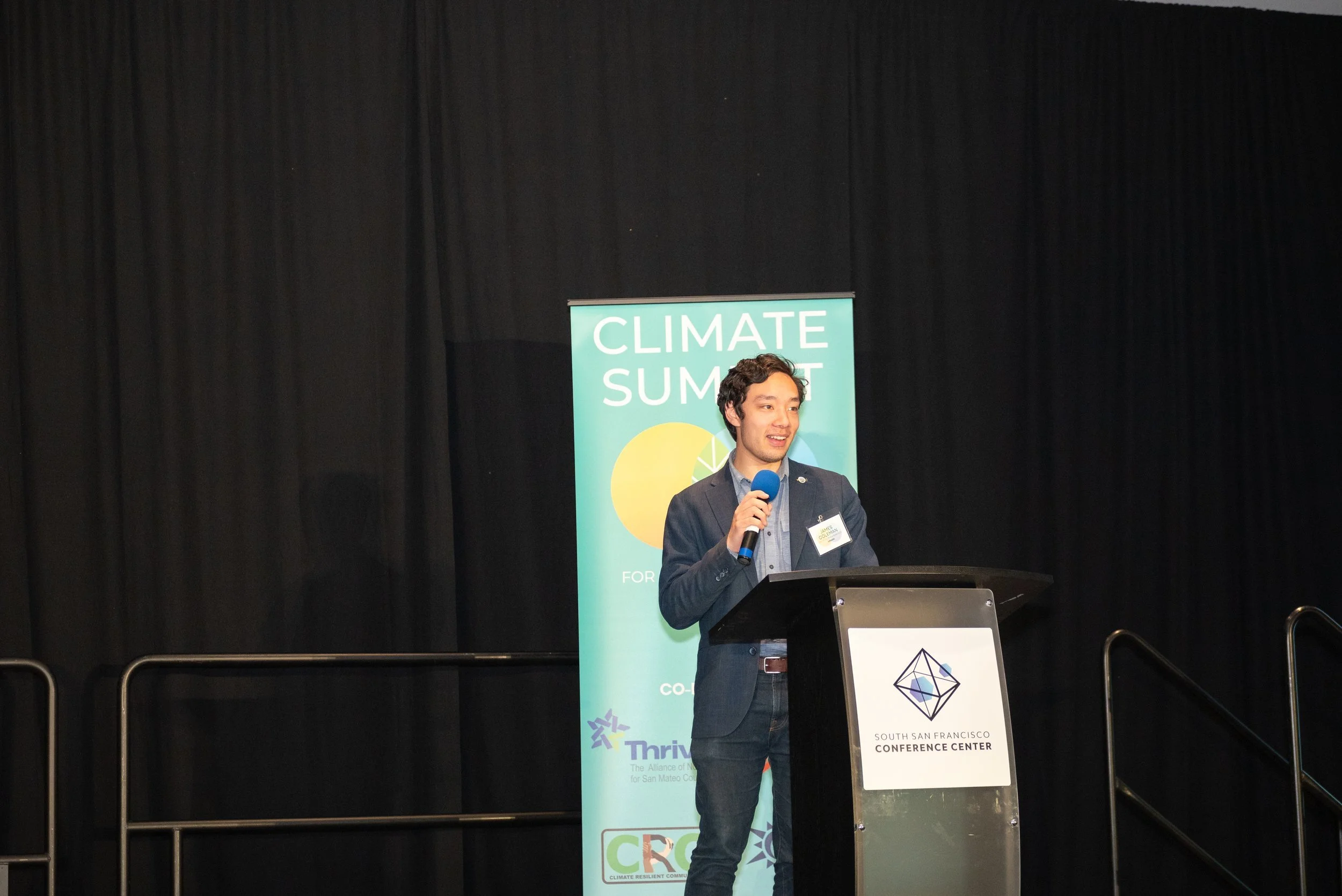 A man giving a speech at a conference, standing behind a podium with the South San Francisco Conference Center emblem. He holds a blue microphone and wears a dark blazer over a light shirt.