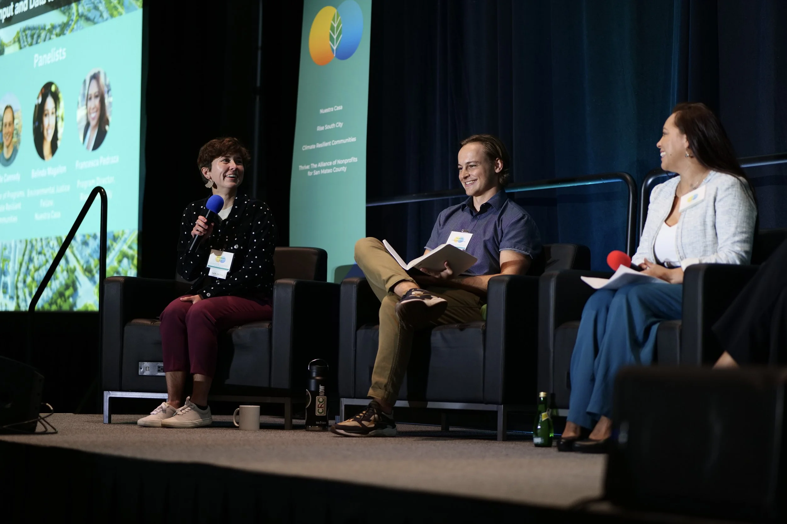 Panel discussion at a conference with three people on stage sitting in black chairs with microphones, a large screen displaying panelist information, and a logo with a leaf on a colorful background.
