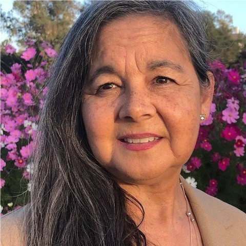 A woman with long gray hair and earrings smiling outdoors with pink flowers in the background.