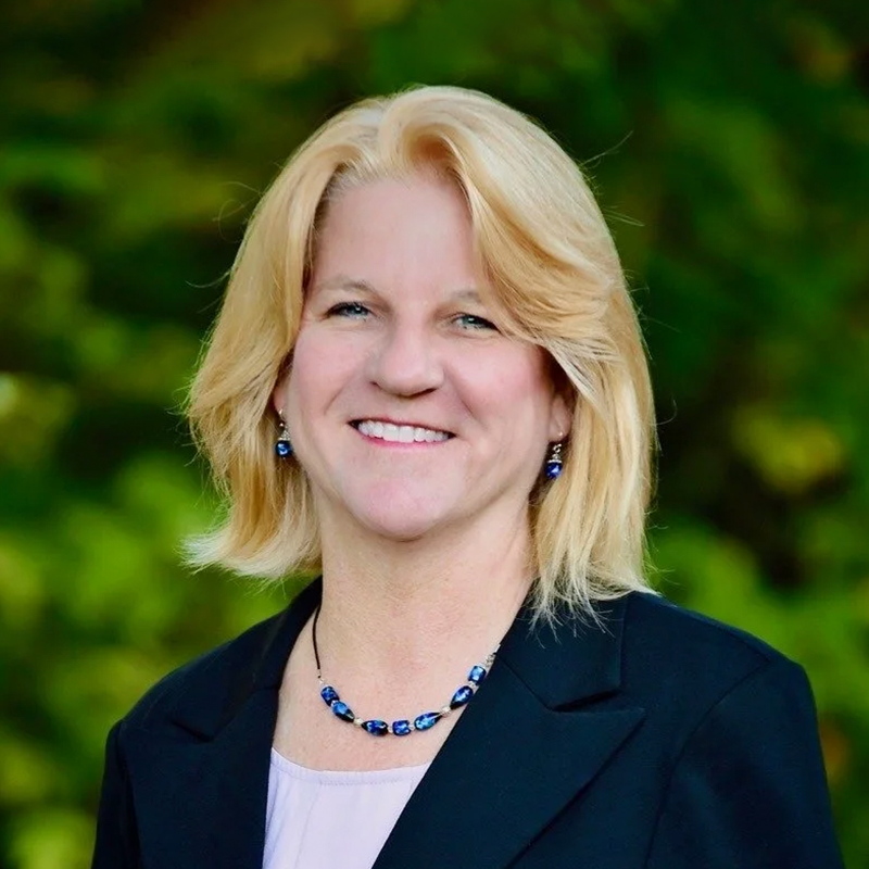 A blonde woman smiling, wearing a black blazer, a light-colored blouse, and blue jewelry, with a green blurred outdoor background.