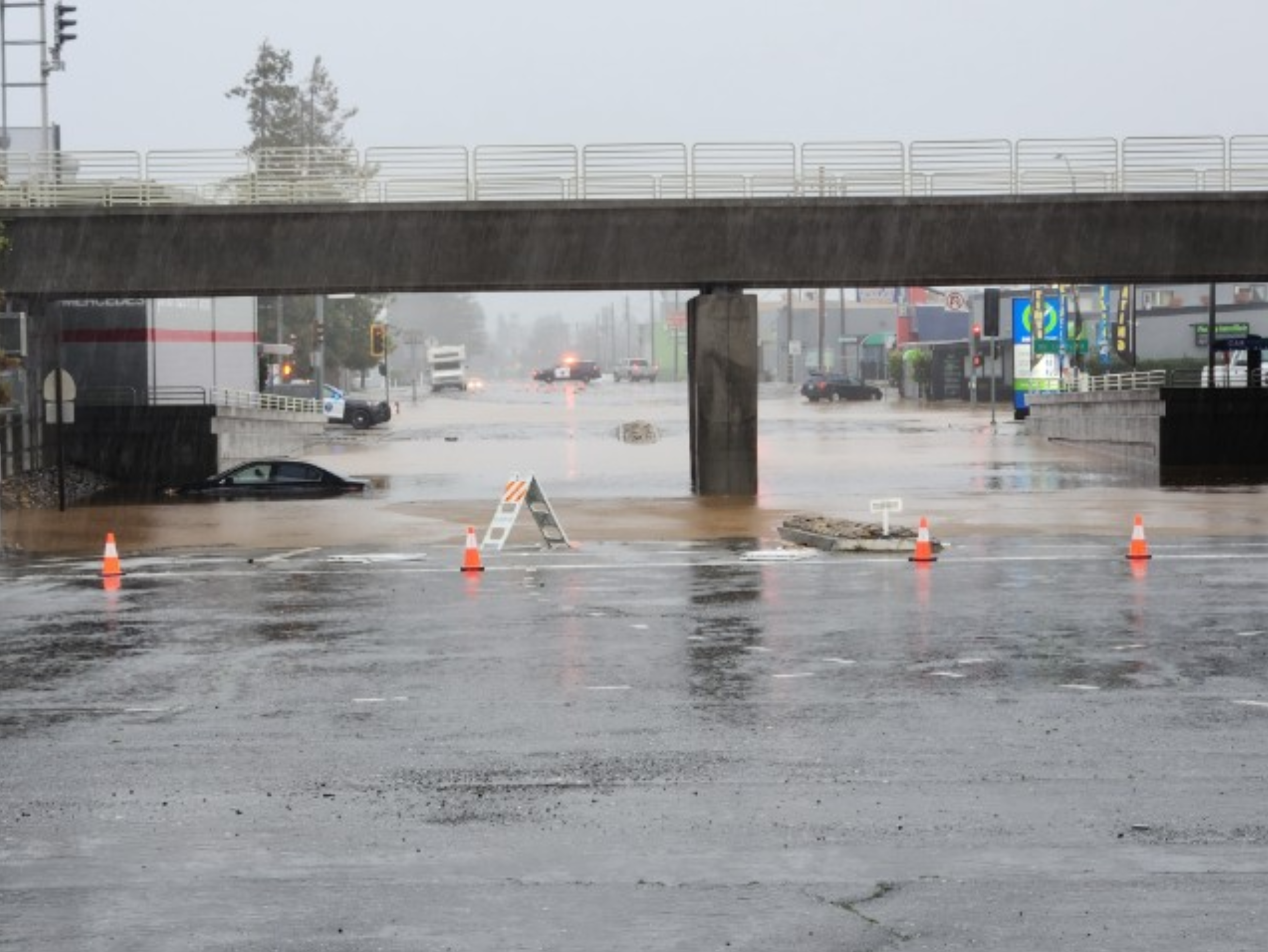 A flooded city street with submerged cars, traffic cones, and an overpass, during heavy rain.