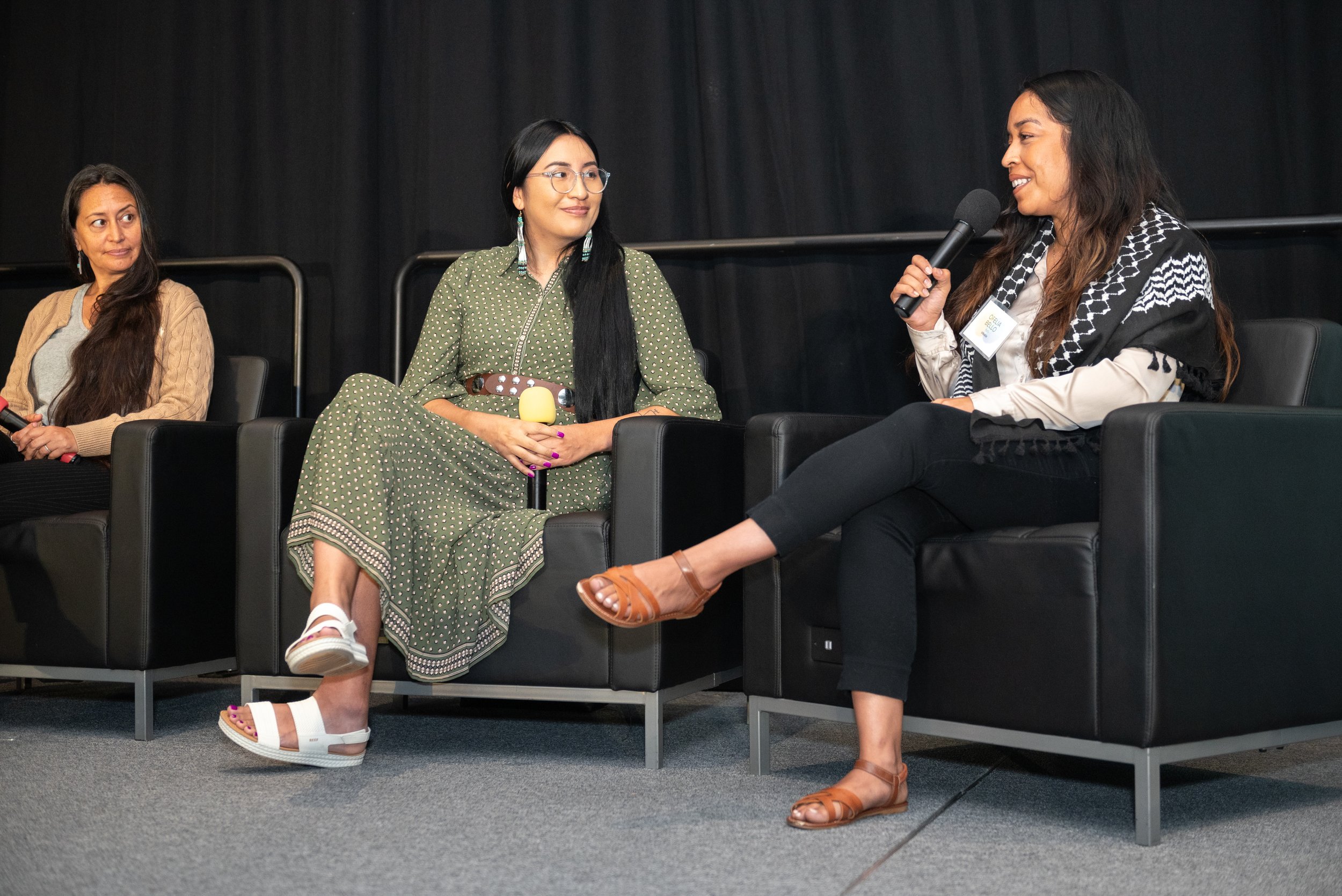 Three women sitting on black armchairs in a panel discussion. The woman on the right is speaking into a microphone, wearing beige shirt, black pants, and brown sandals. The woman in the middle wears a green polka-dot dress and glasses, holding a yell