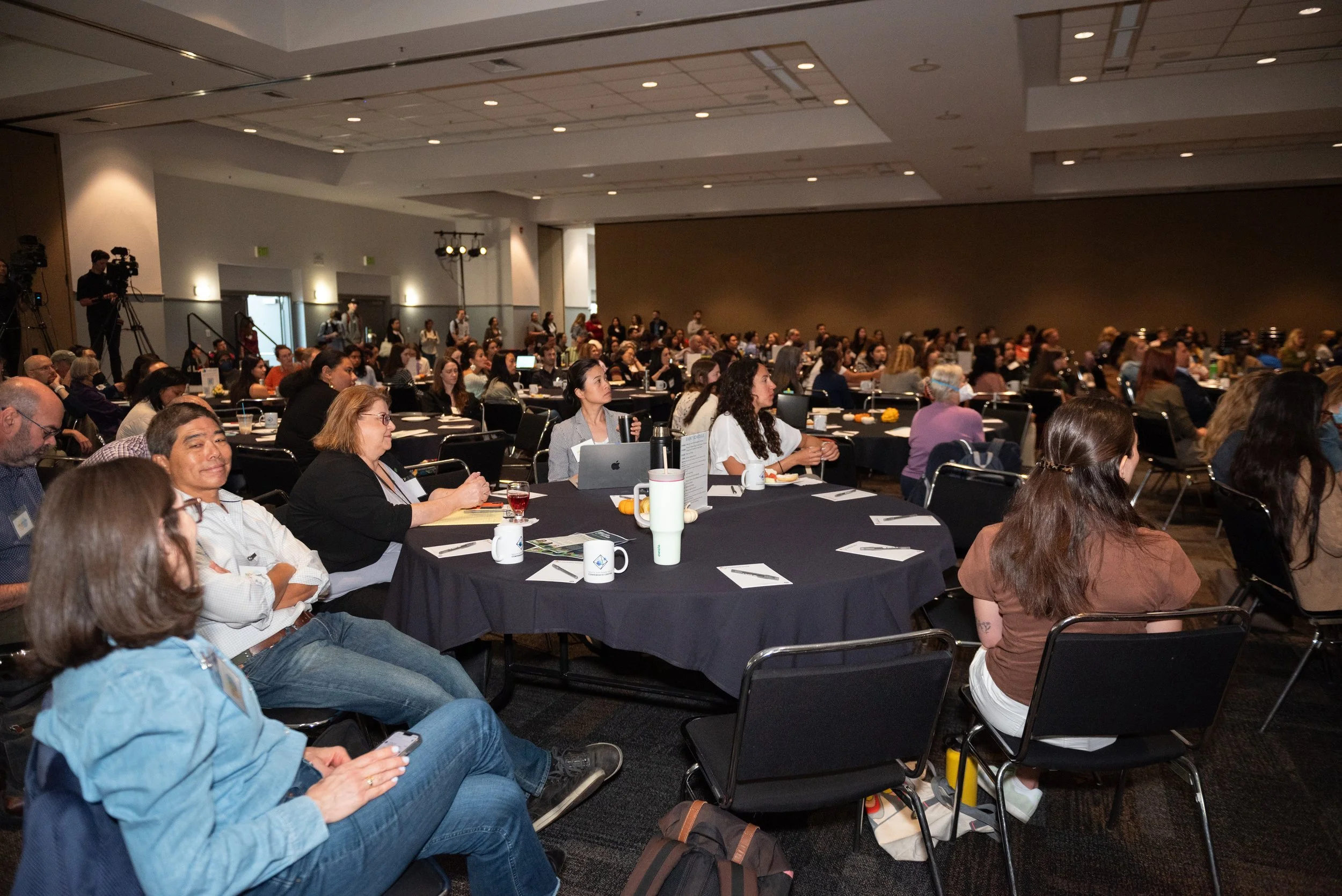 A large conference room filled with many attendees seated at round tables, listening to a presentation or speaker at the front of the room.