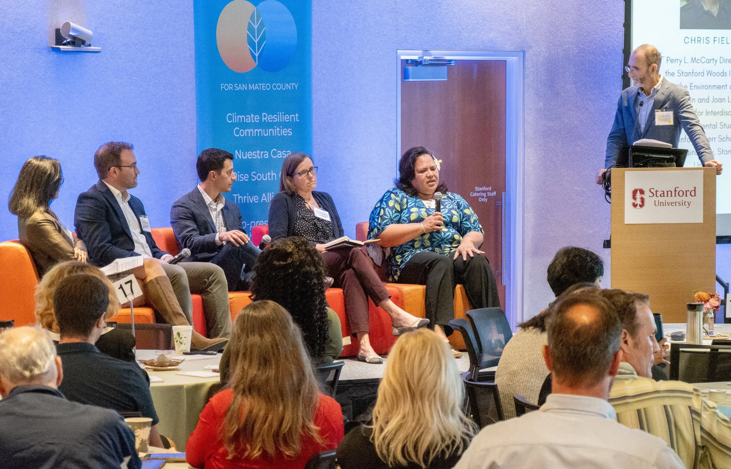 Panel discussion at Stanford University event with speakers seated on a stage. One woman is speaking into a microphone. Audience members are seated at tables in front of the stage.