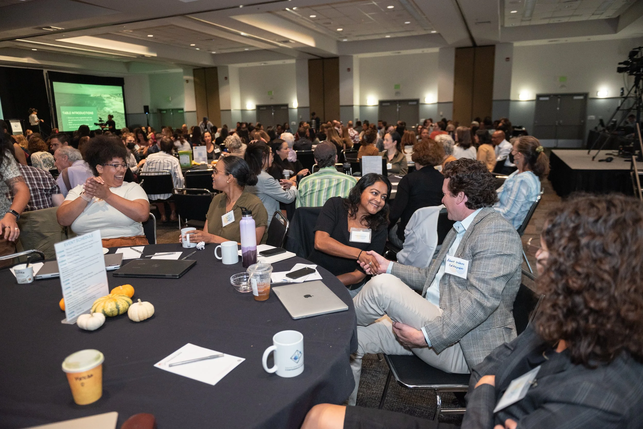 A diverse group of people sitting at round tables in a conference room, engaged in conversation. The tables have laptops, coffee cups, and small decorative pumpkins. The room is filled with attendees, and a stage with a large screen is visible in the