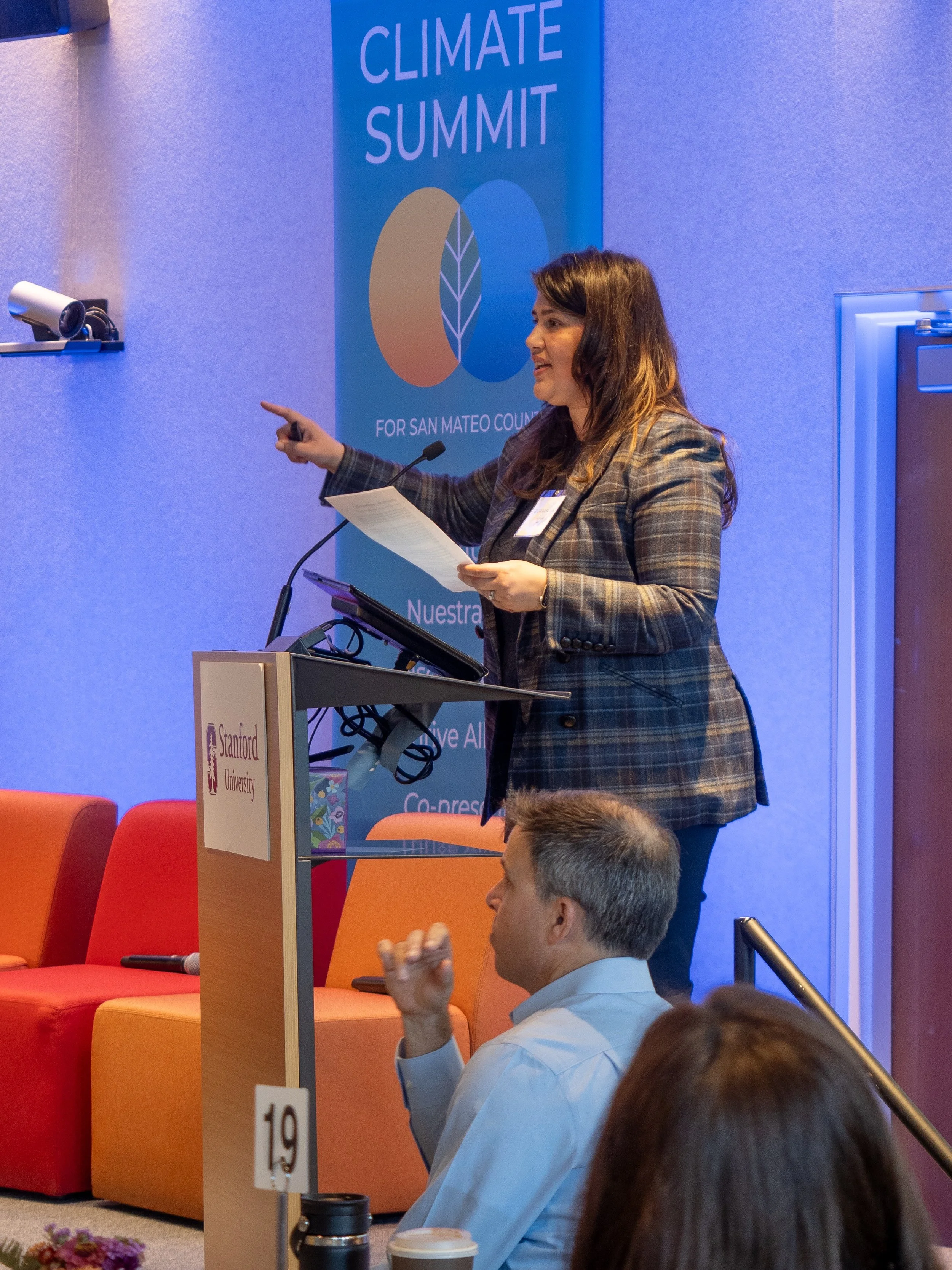 Woman speaking at a podium with Stanford University logo during a climate summit event, with an audience member listening attentively.