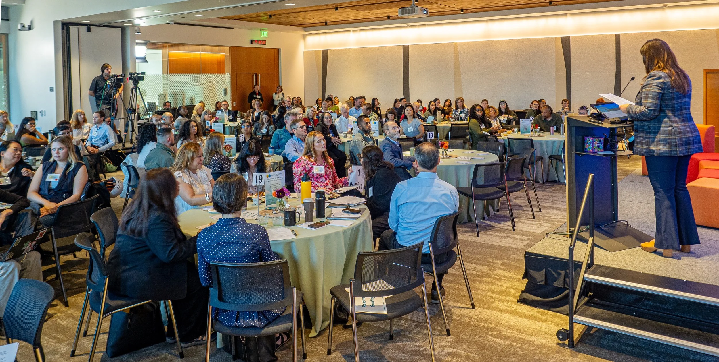 A woman wearing a plaid blazer is giving a presentation at a conference, standing on a small stage with a lectern, addressing an audience seated at round tables.