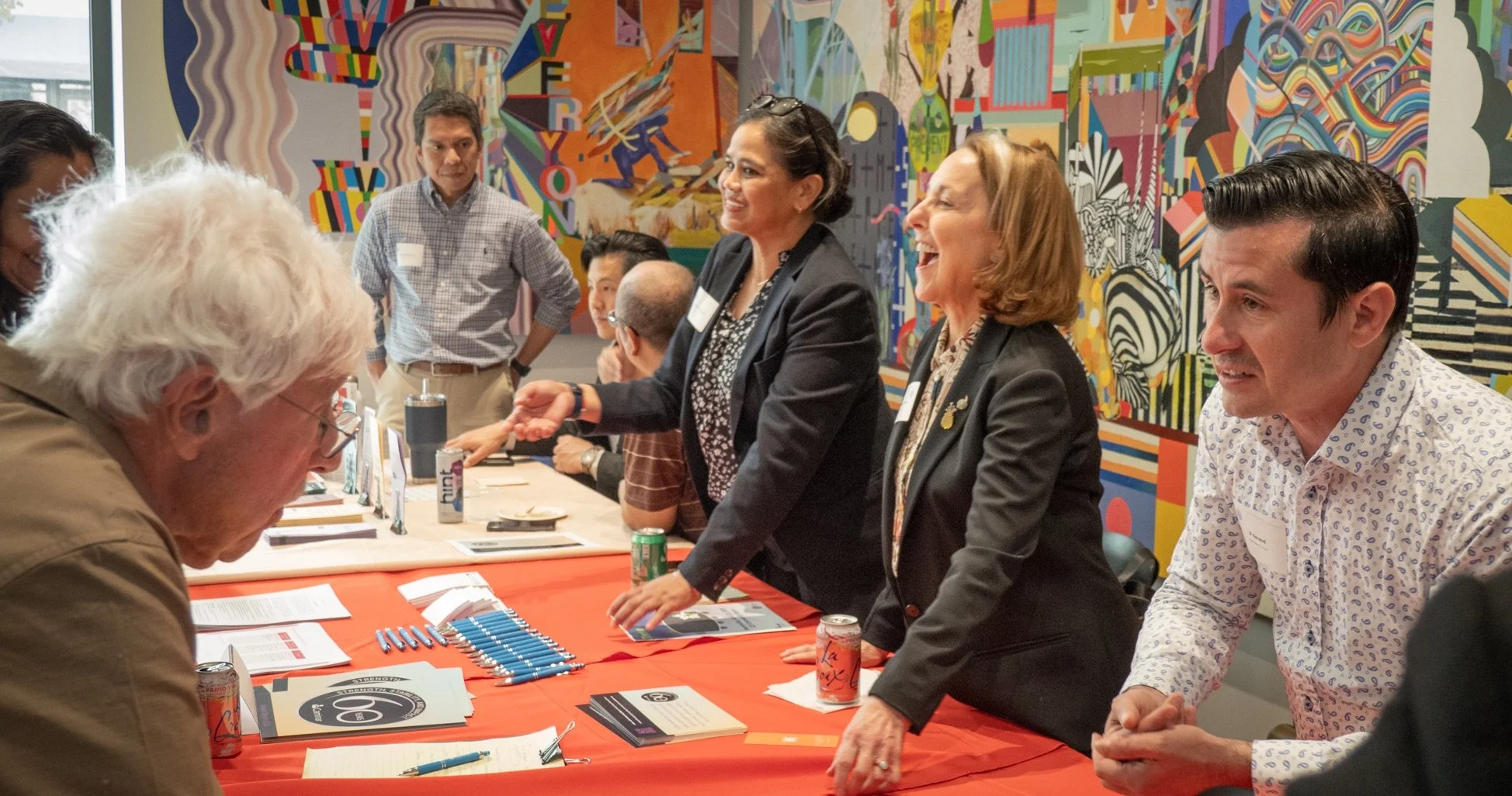 A diverse group of business professionals are engaged in a meeting or discussion around a table, with some smiling and talking. The background features a colorful, abstract mural.