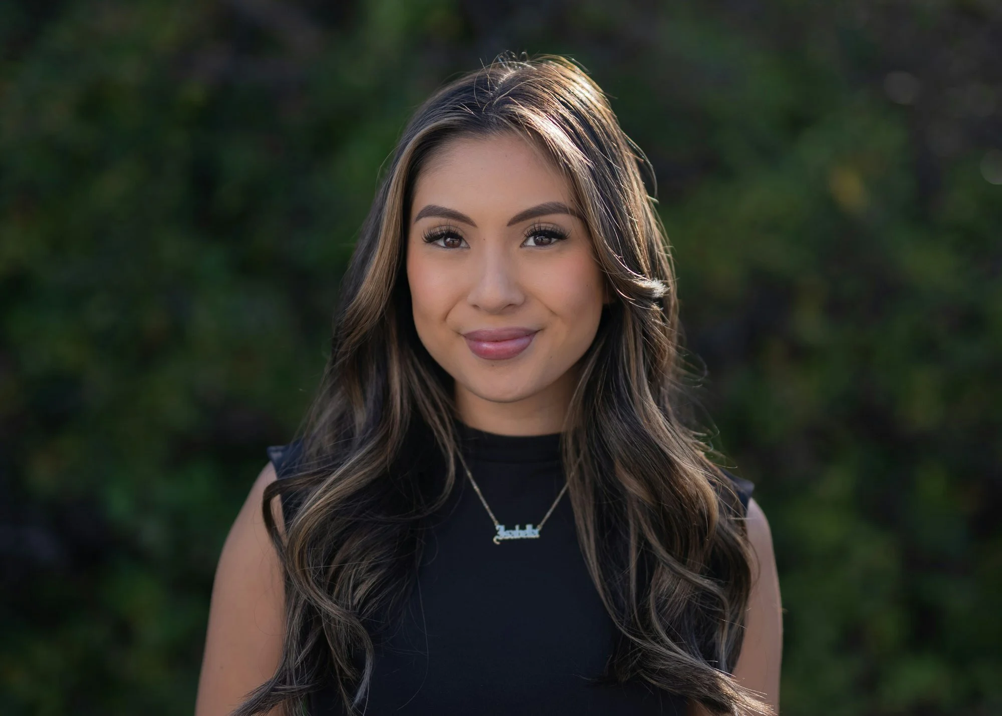 A young woman with long wavy hair smiling outdoors, wearing a black sleeveless top and a necklace with her name on it.