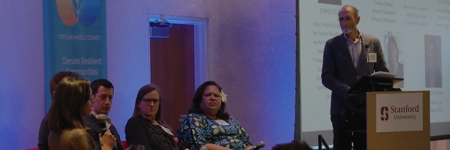A panel of four people sitting at a table while a man stands at a podium giving a presentation at Stanford University. The background features a blue banner with text about climate resilient communities and a large projection screen.