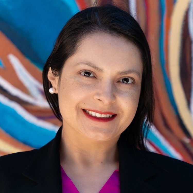 A woman with black hair, wearing pearl earrings and a black blazer over a pink top, standing in front of a colorful abstract background.