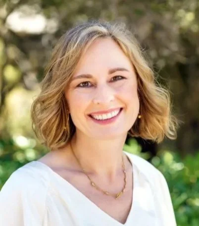 A smiling woman with short blonde hair standing outdoors with greenery in the background.