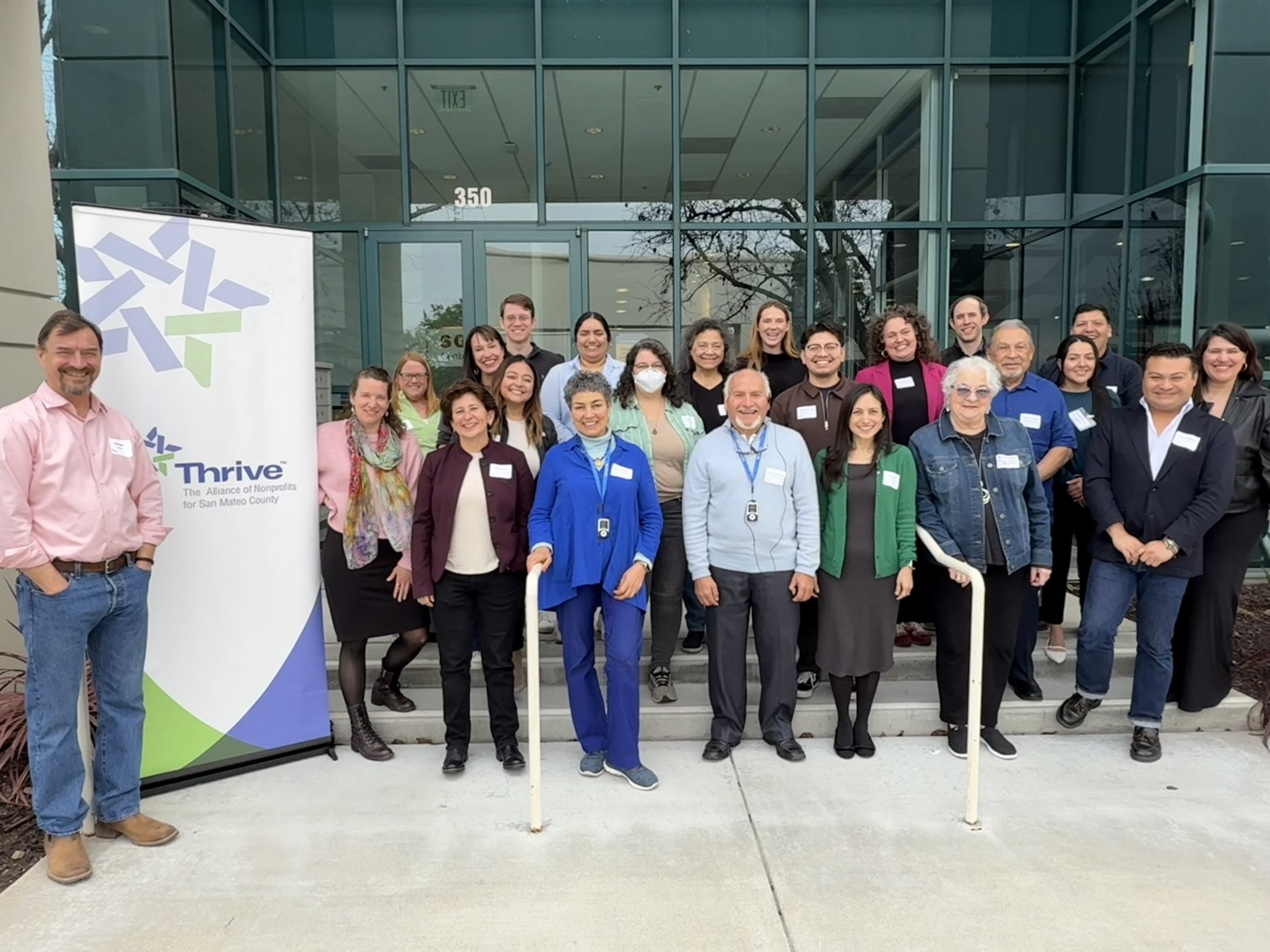 Group of diverse people standing outside a modern building with glass doors, posing for a photo near a Thrive banner.