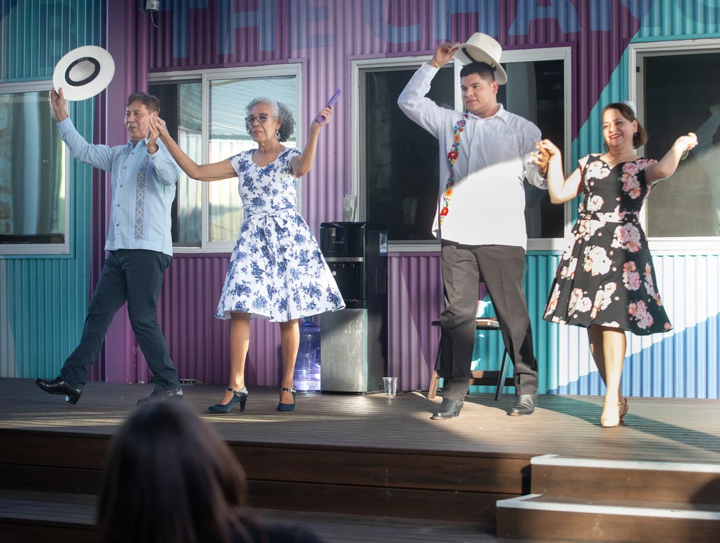 Four people dancing on a stage with a colorful background, two men and two women, dressed in vintage-style clothing, holding cowboy hats and dancing happily.