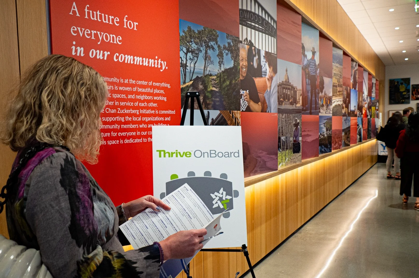 A woman with curly blond hair reading a pamphlet at an indoor community event with a large red wall display that reads 'A future for everyone in our community' and features photographs of local scenery and people.