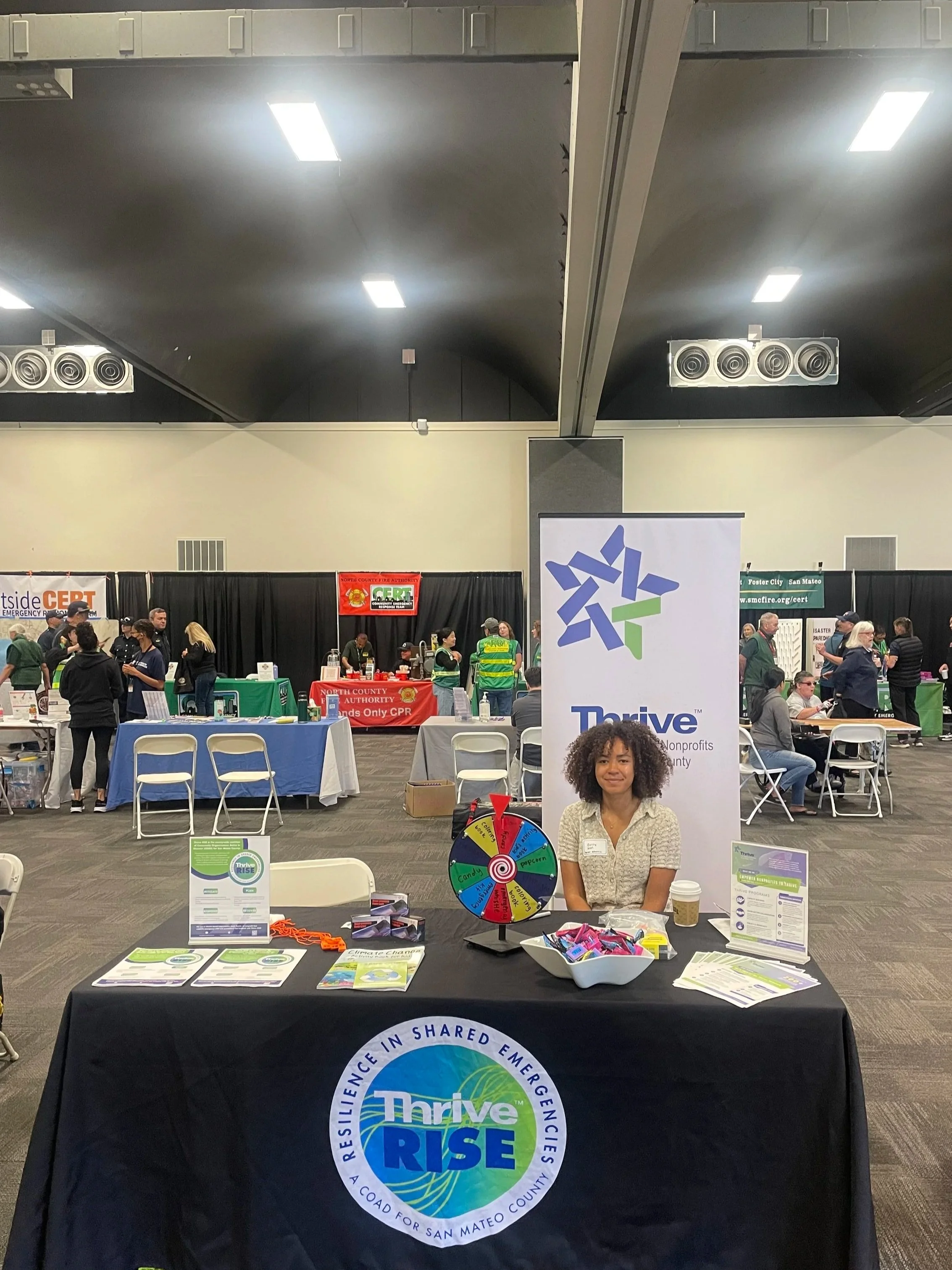 A woman sitting at a booth with a Thrive RISE banner at an indoor event or conference. The booth has a spinning wheel and various informational materials. In the background, there are other booths and people walking around.