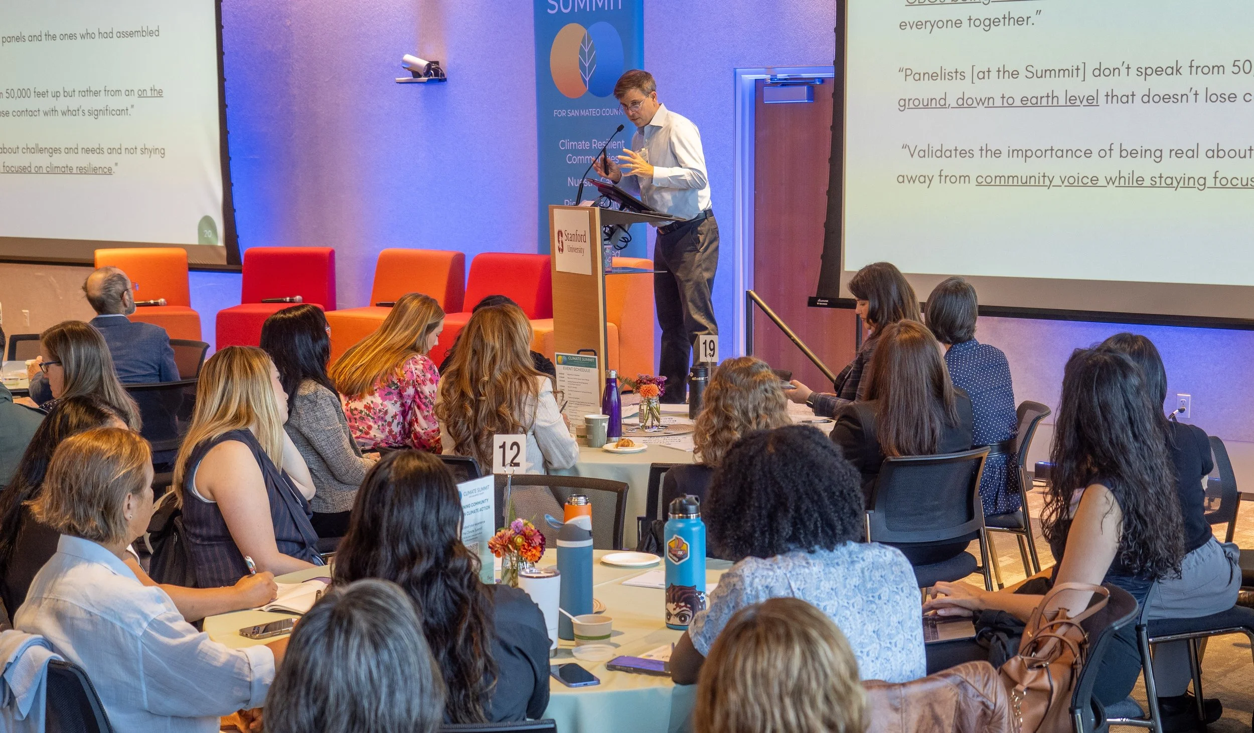 A speaker giving a presentation at Stanford University during a climate summit. Audience members are seated at round tables, taking notes and listening attentively, with large screens displaying presentation slides behind the speaker.