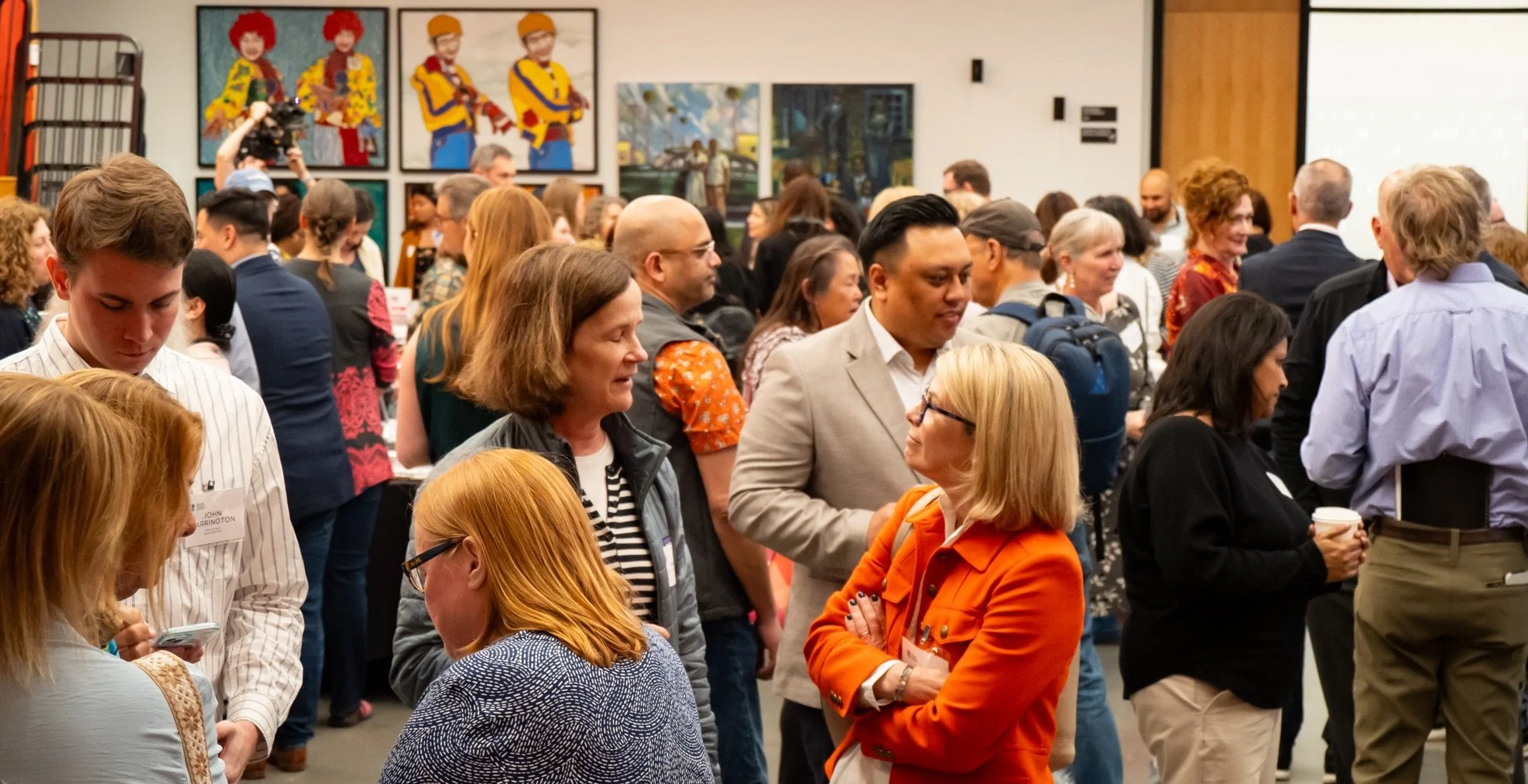 Crowd of diverse individuals attending a conference or seminar in an art gallery setting, engaging in conversations, with colorful paintings on the walls.