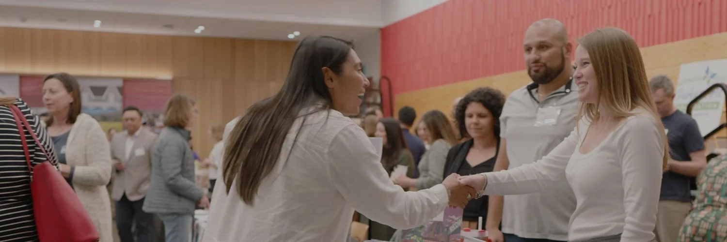Two women shaking hands at a busy indoor event, with several other attendees visible in the background.