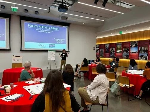 A woman presenting at a workshop titled 'Policy Affinity Group' in a conference room with attendees seated at red-covered tables.