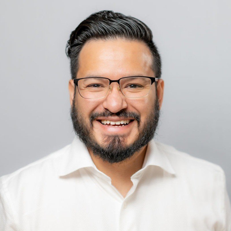 Smiling man with glasses and beard wearing a white shirt against a plain backdrop.