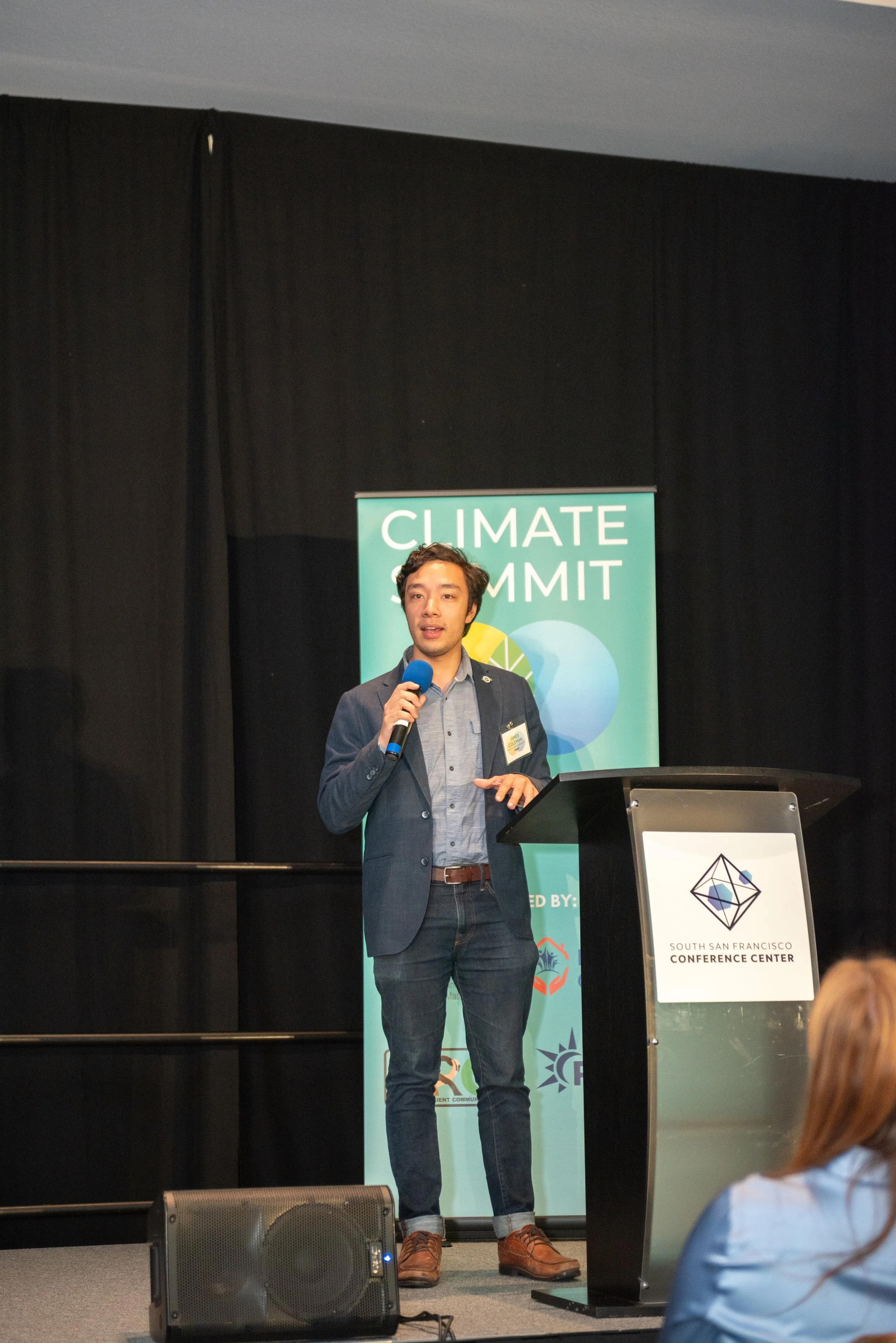 A man in a blazer speaking into a blue microphone at a Climate Summit in the South San Francisco Conference Center.