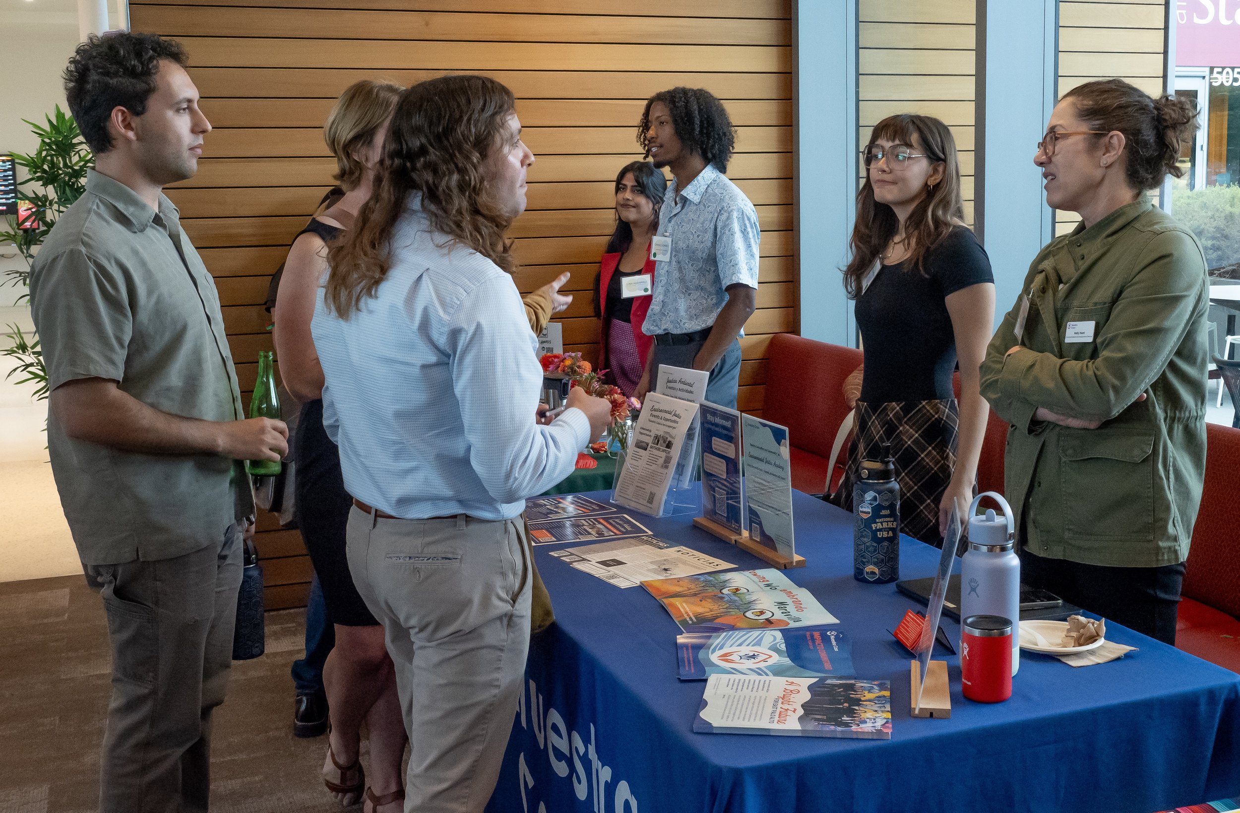 Group of people at an indoor informational booth, engaging in conversation. The booth display includes pamphlets, brochures, and signs, with a blue tablecloth and background wooden paneling.