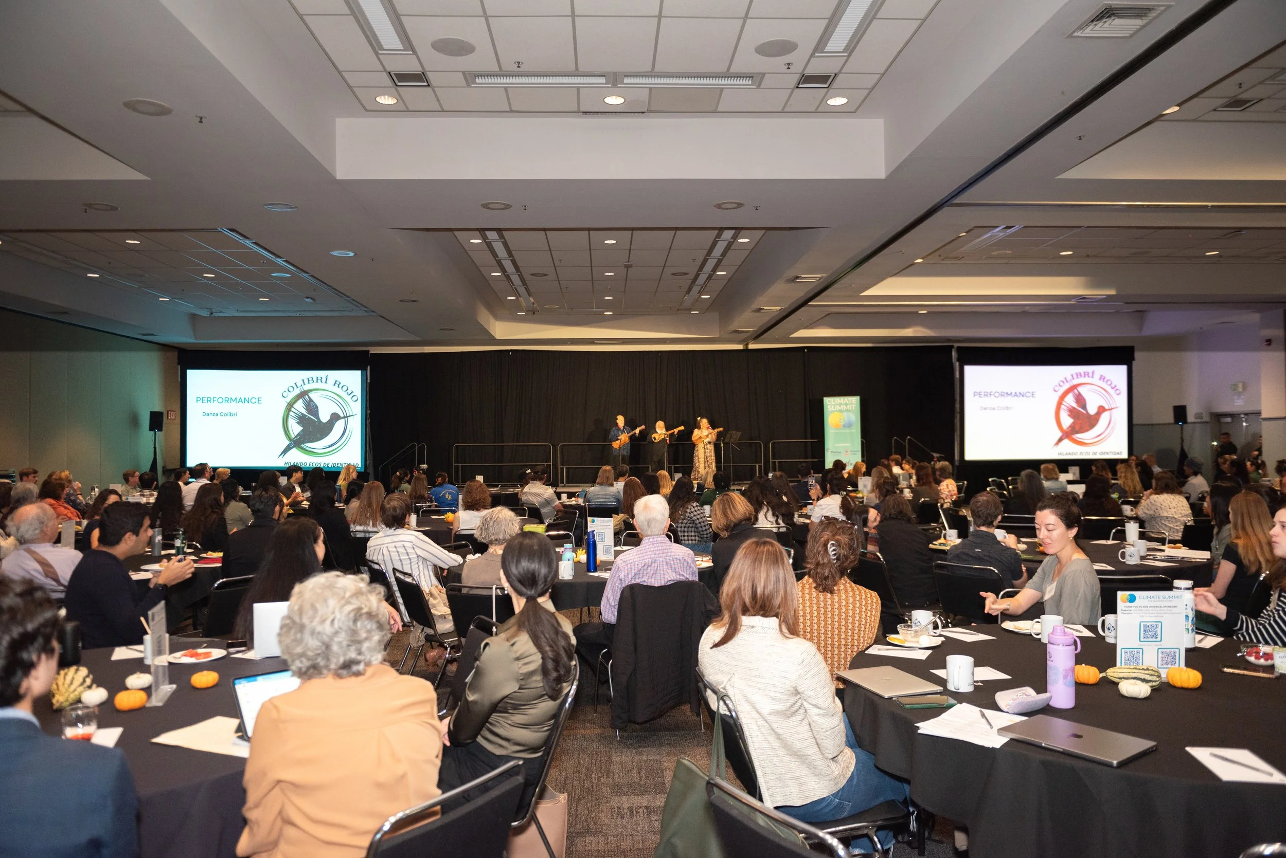 Audience attending a performance on stage at conference with two screens displaying 'Performance Danza Colibrí Rojo' and 'Hilando ecos del silencio' in a large conference hall.