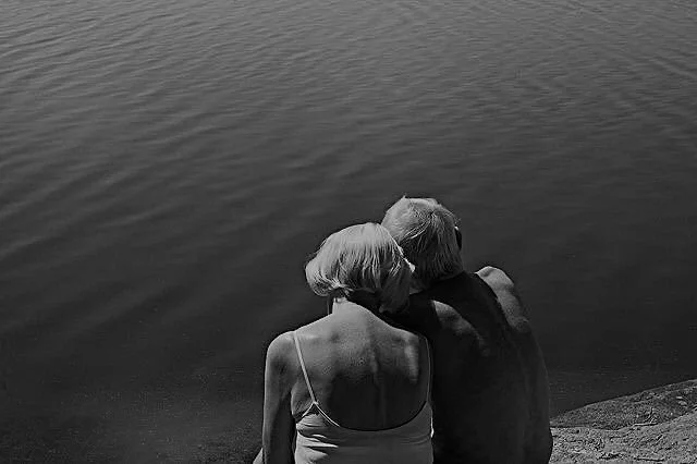An elderly couple sitting by a body of water, sharing a quiet moment with their heads touching.
