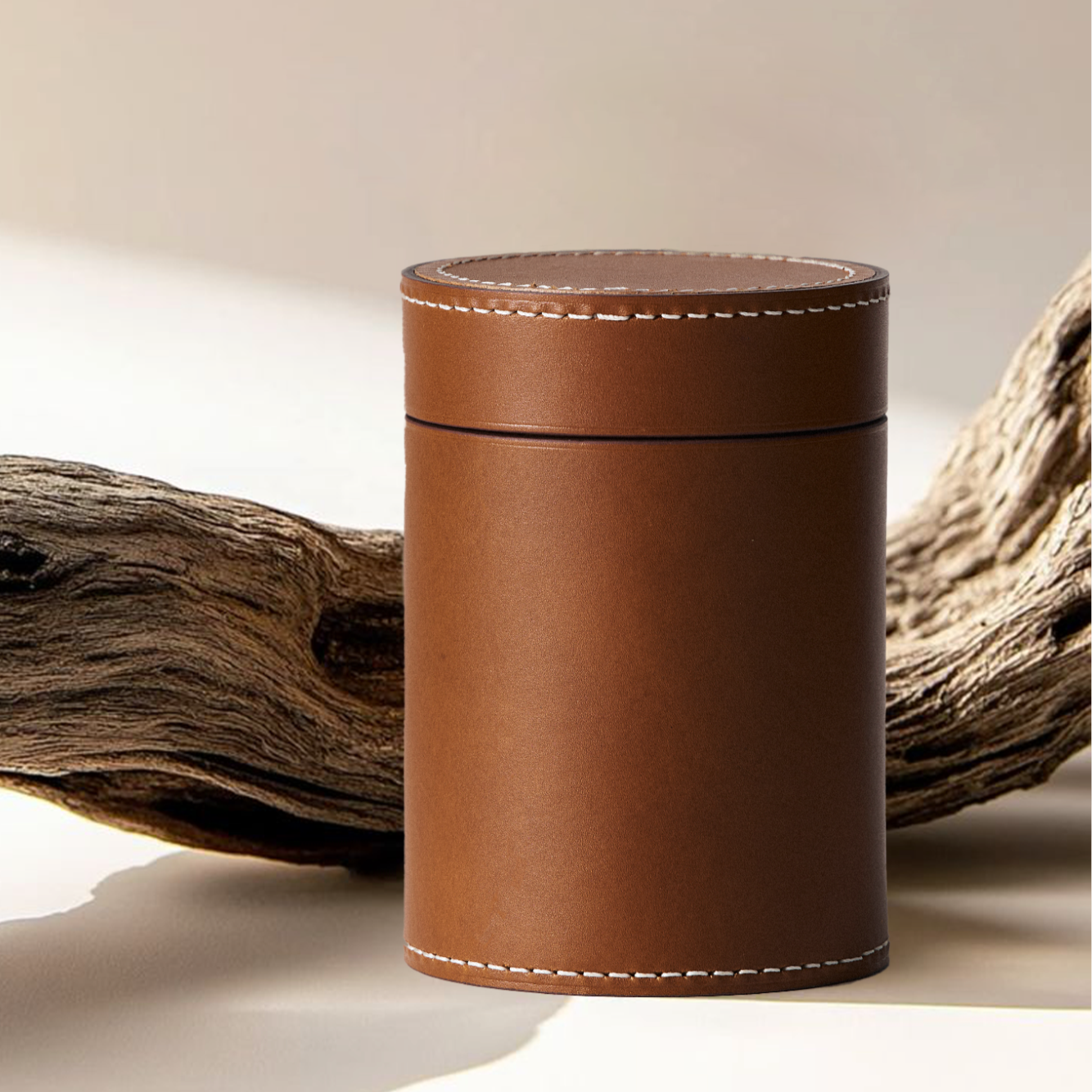 Cognac leather cremation urn with cream stitching, displayed beside driftwood on a pale surface.